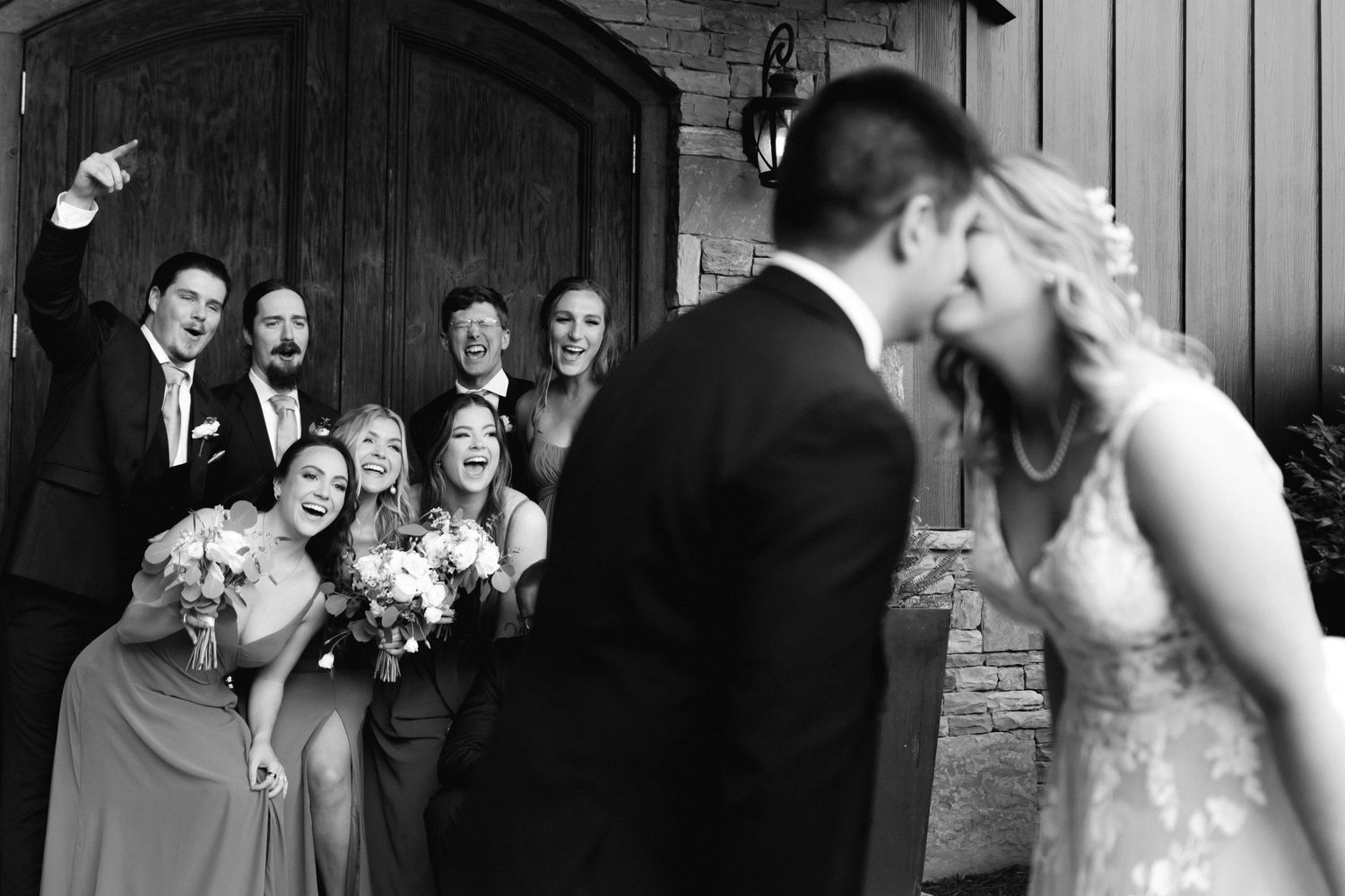 Wedding party reacts with joy as newlyweds share a kiss in front of wooden doors. Blue mountain vineyard wedding photographer in Dahlonega.