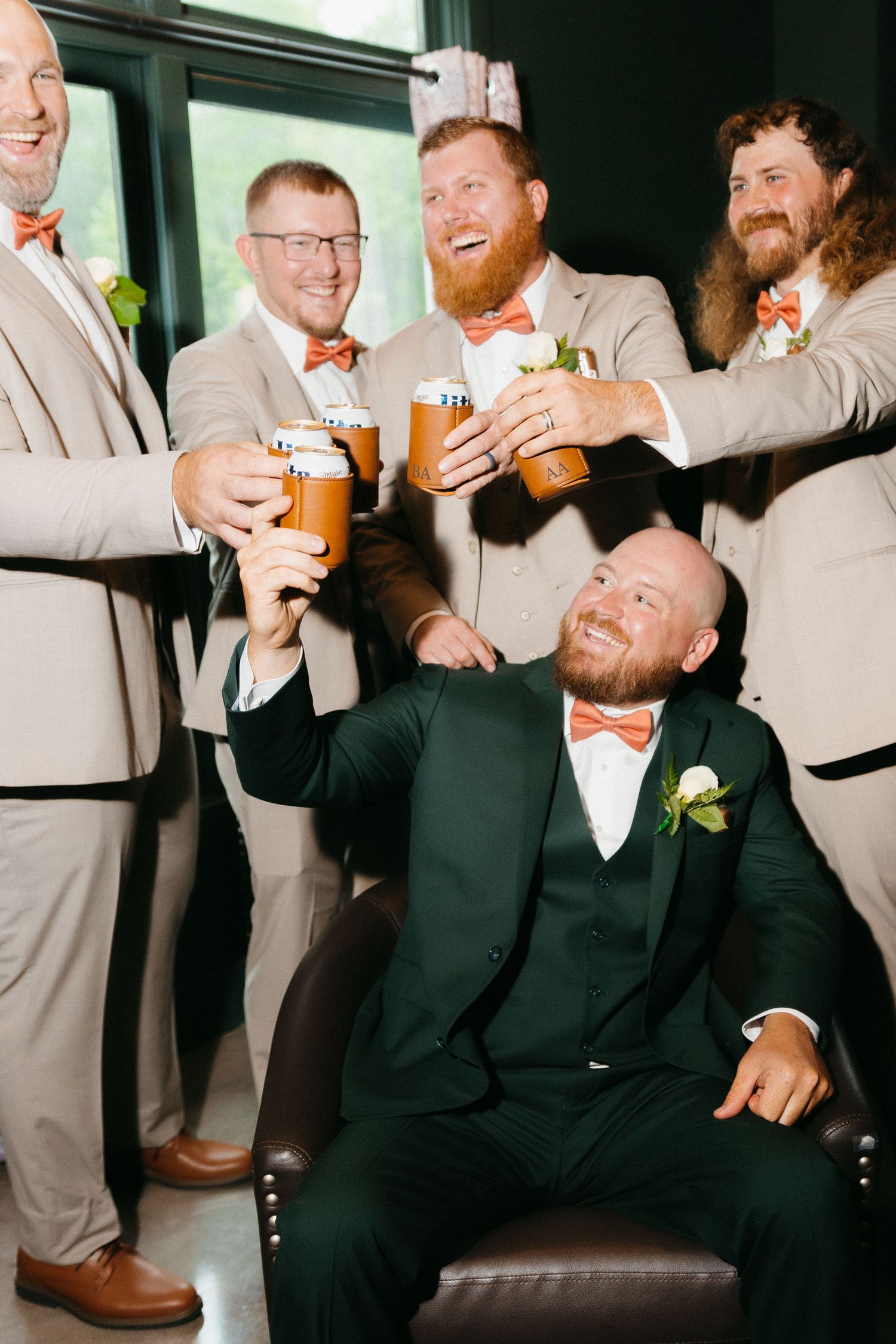 Groomsmen in tan suits and orange bowties toast with drinks while celebrating. Wedding photographer in Northeast Georgia.