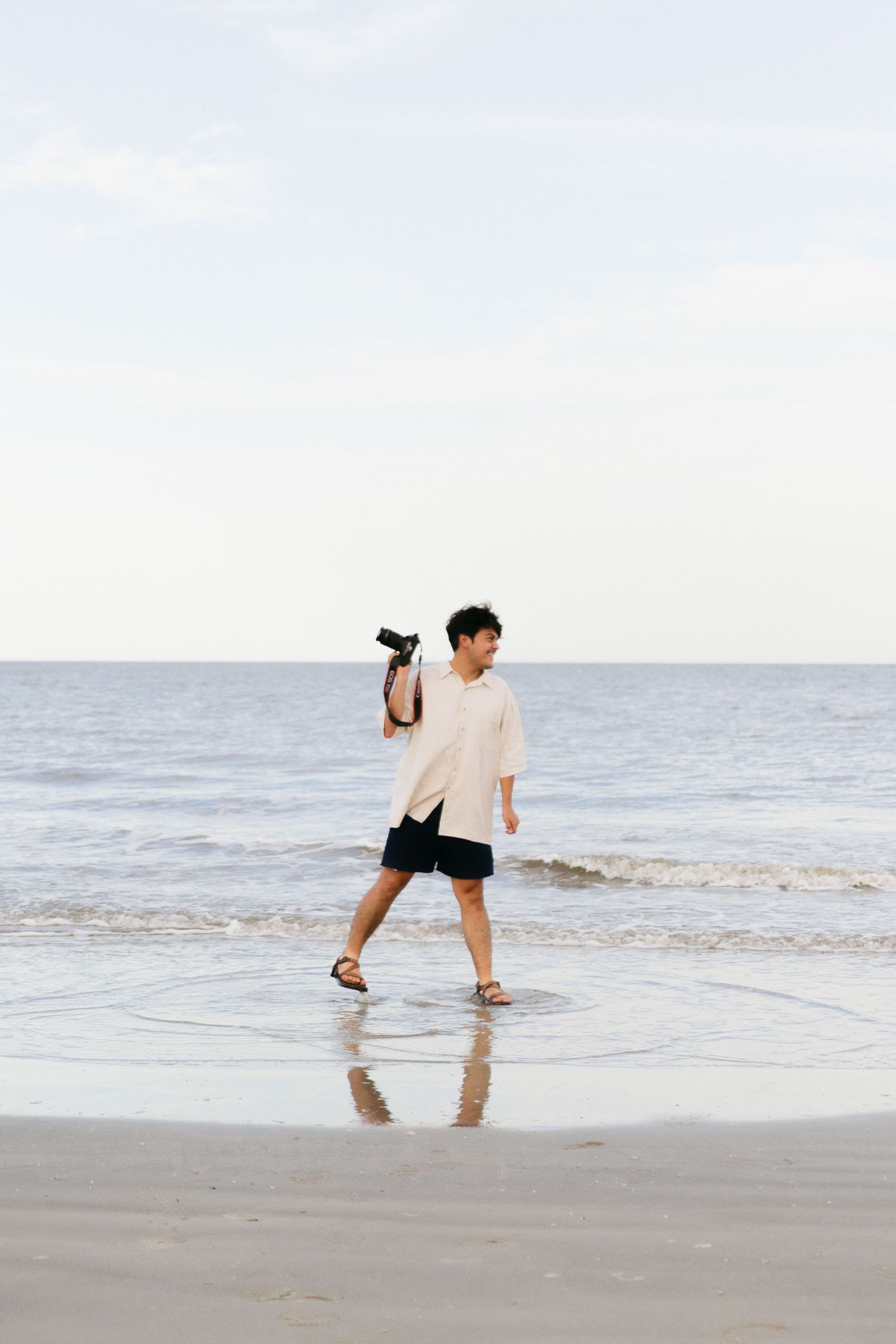 Person in casual attire walking along the beach shoreline holding a camera. Jeckll Island wedding photographer.