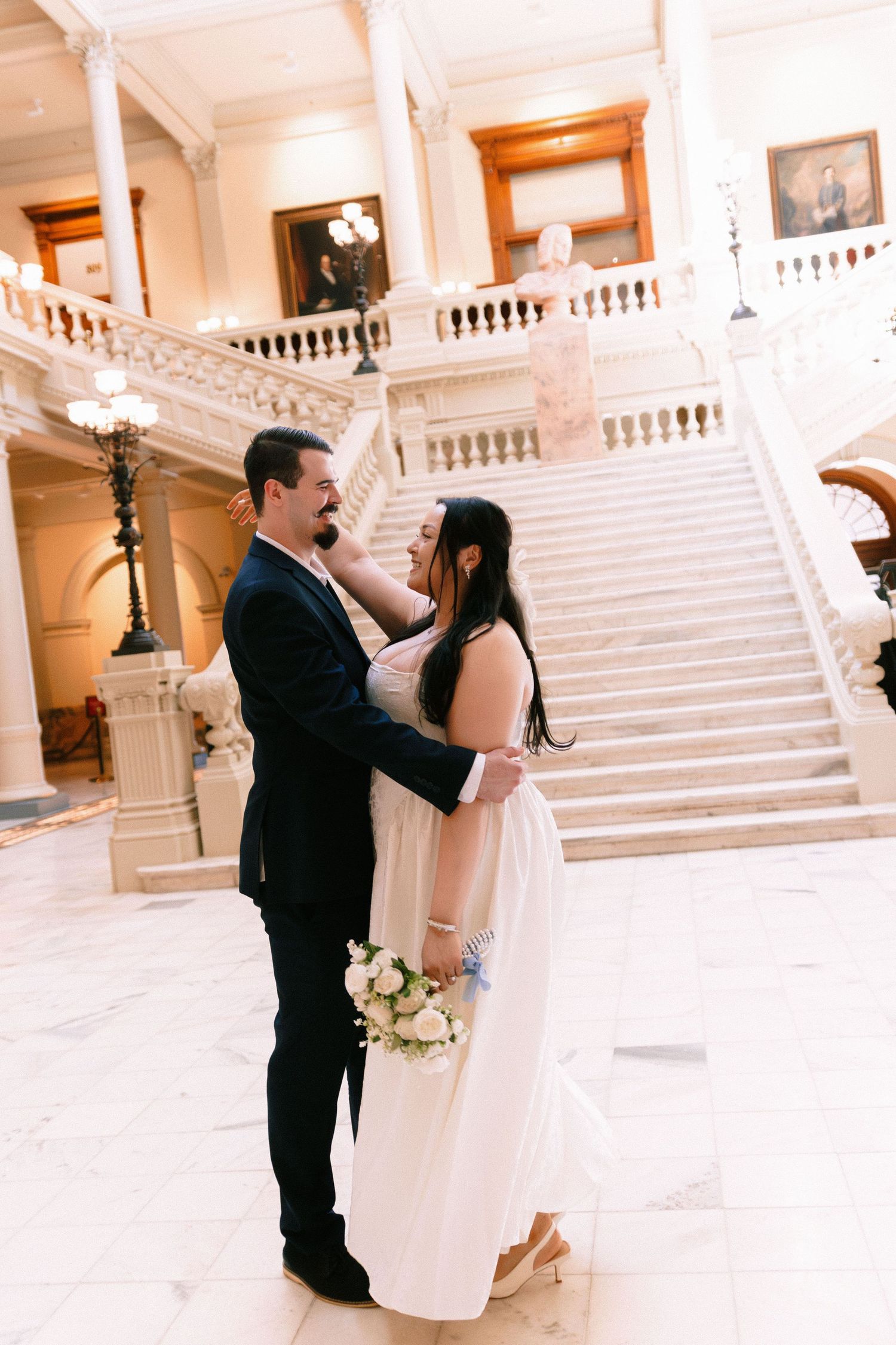 A romantic wedding photo of a couple embracing in front of an elegant white marble staircase in a grand building. Georgia photographer.
