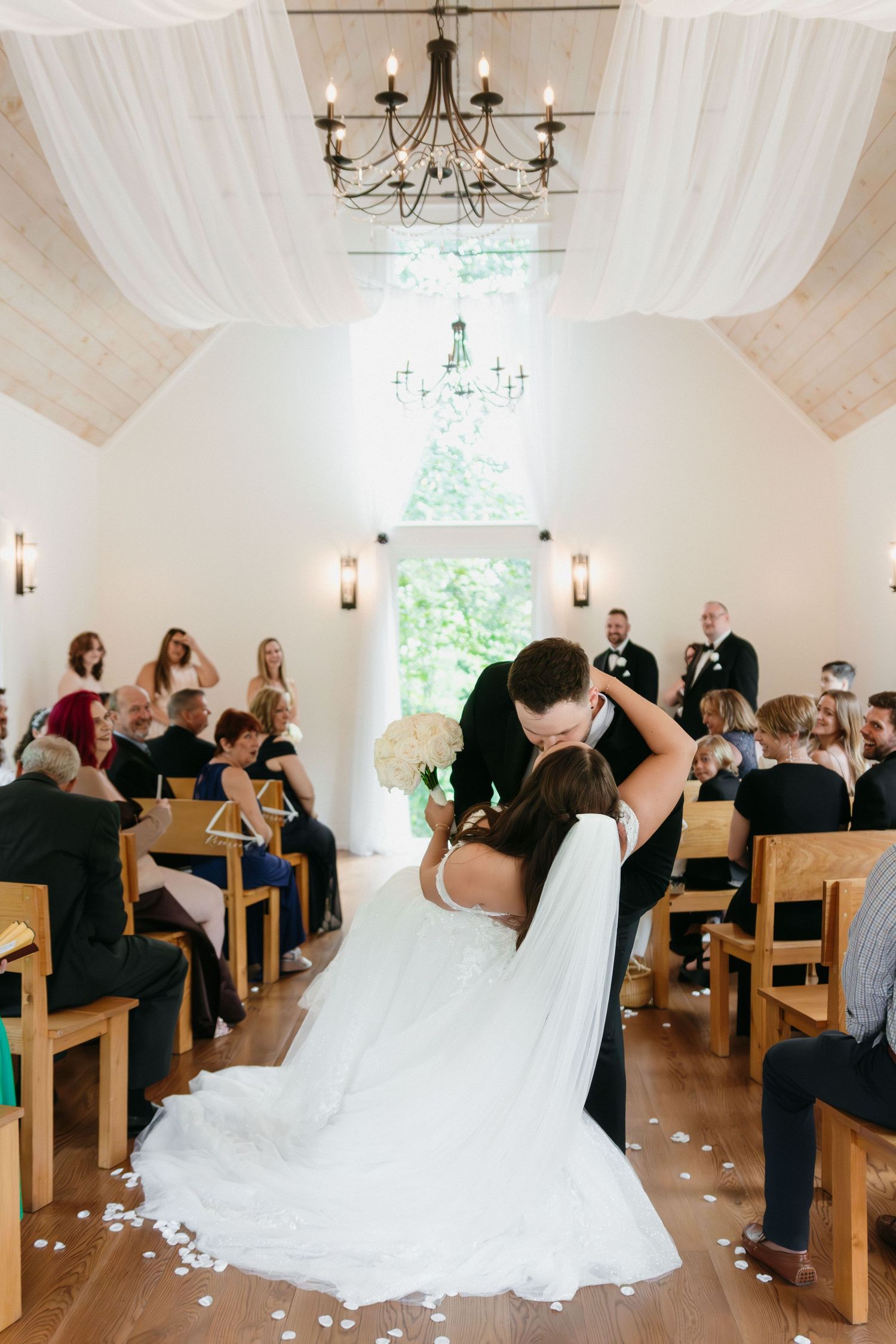 Joyful wedding ceremony in a rustic white chapel with wooden pews and scattered flower petals down the aisle.