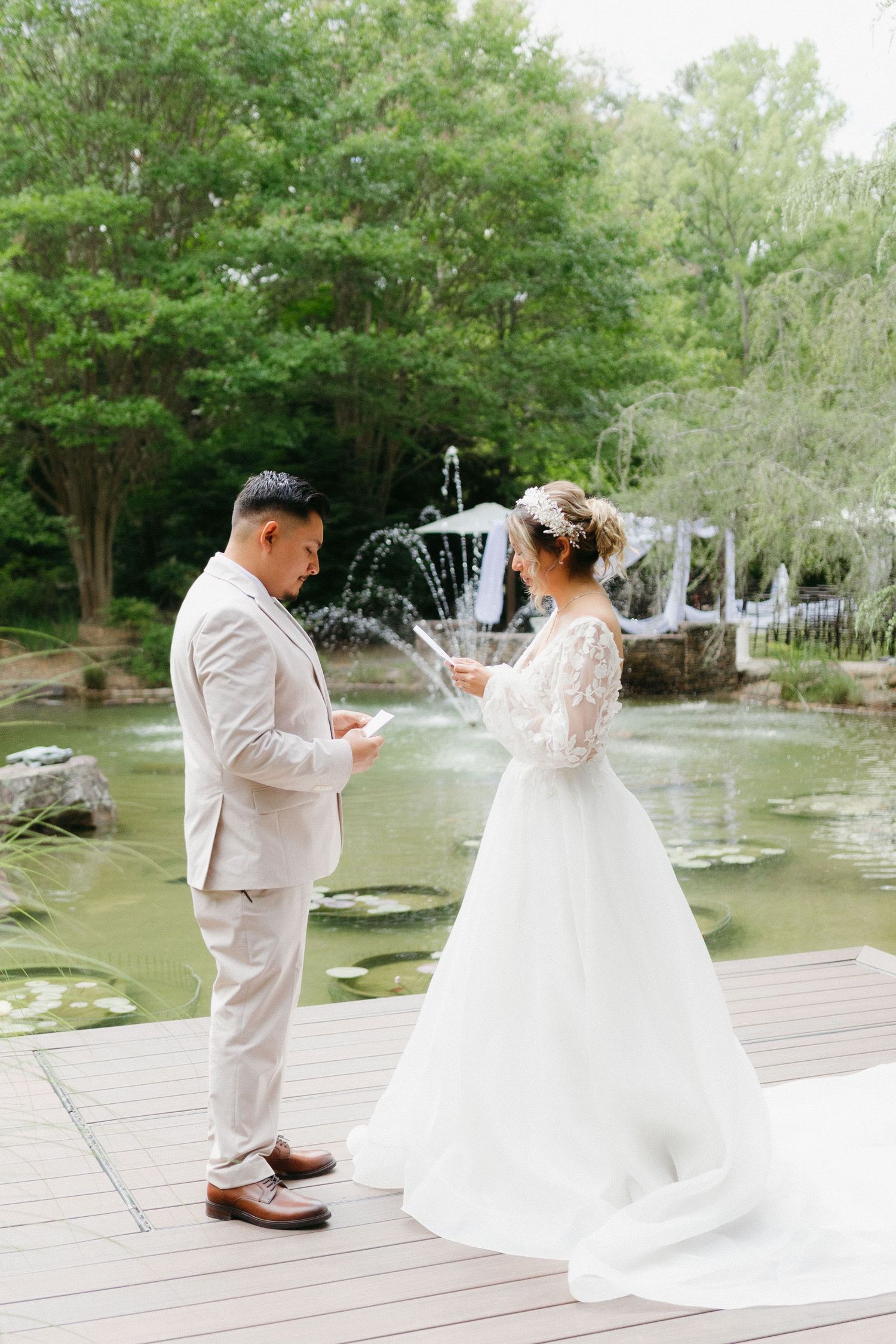 A couple exchanges vows during their wedding ceremony on a wooden deck beside a fountain and pond in a garden setting.