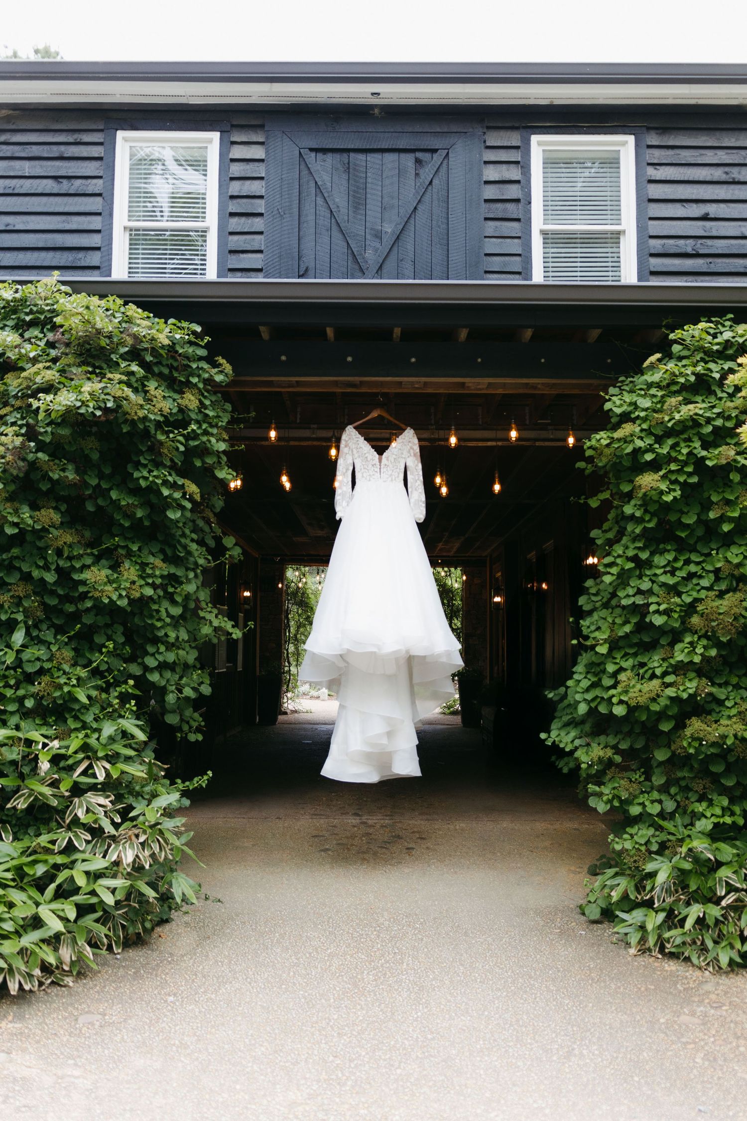 A white wedding dress hangs in a doorway of a navy blue barn-style building with string lights. Cumming, Georgia wedding photographer.
