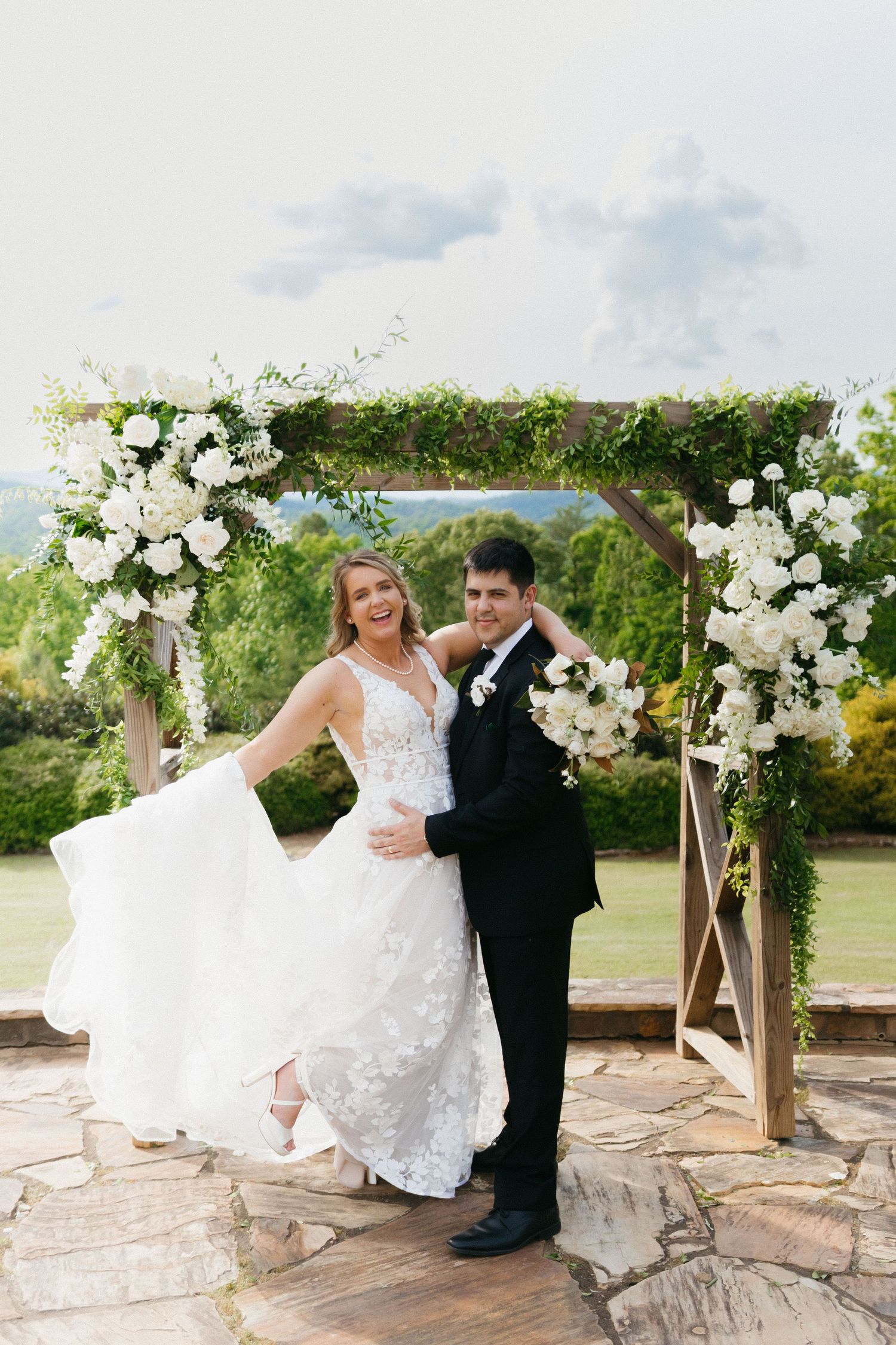 A couple poses under a floral arch with white roses at an outdoor wedding venue. Blue mountain Vineyard wedding photographer in Dahlonega.