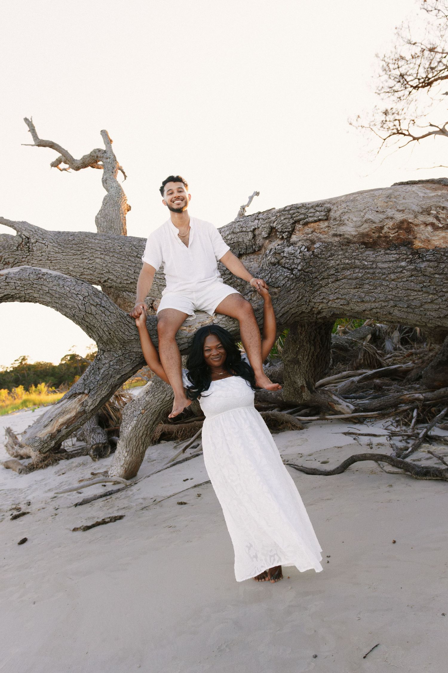 Couple poses playfully on driftwood log at beach during sunset photoshoot. Jekyll island elopement photographer.