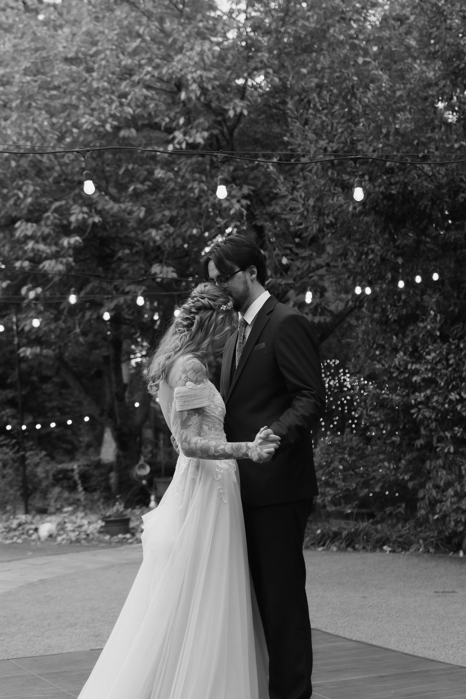 A romantic black and white photo of a couple dancing at their wedding reception with string lights in the background.