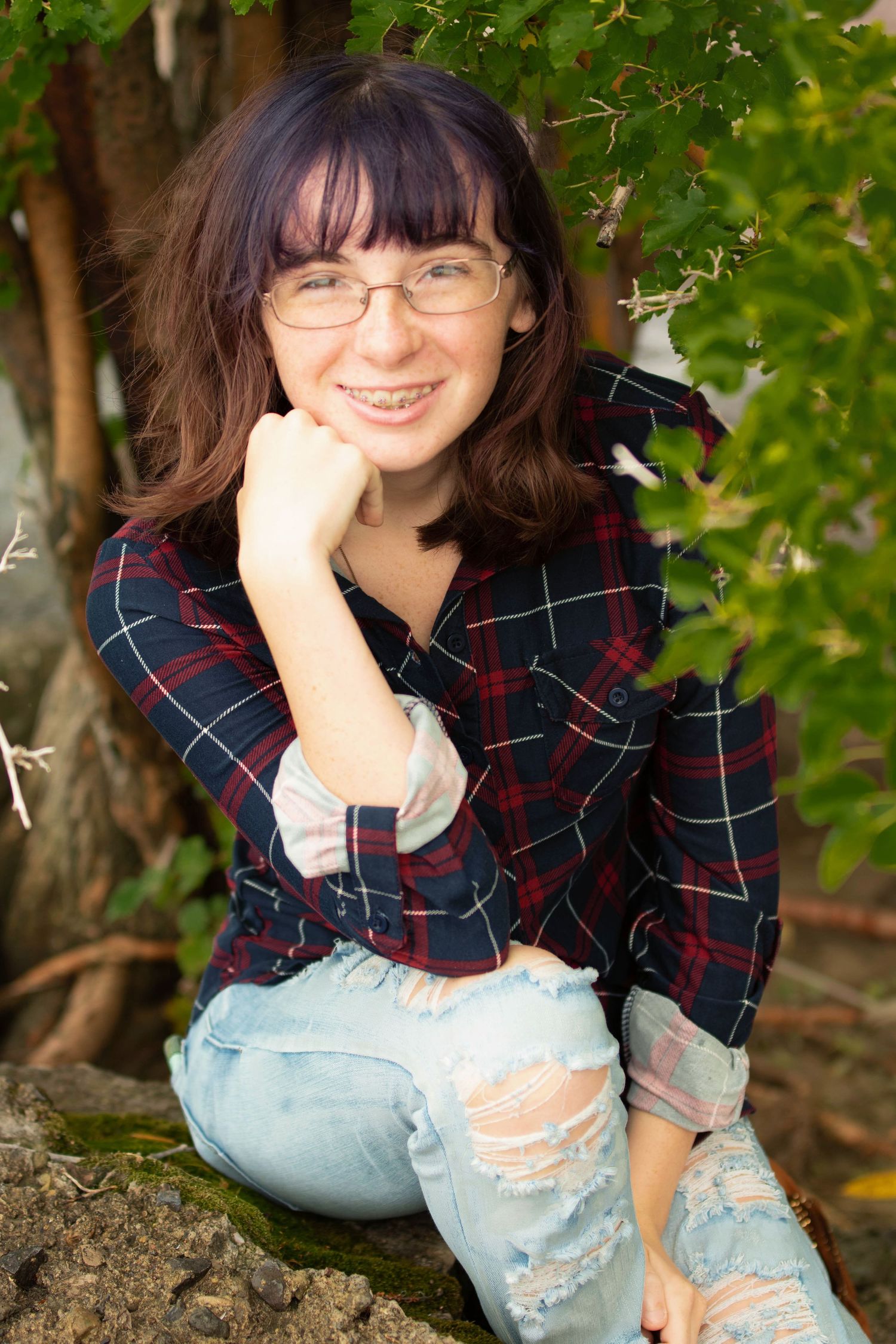 Person in plaid shirt and ripped jeans poses casually on rocks outdoors.