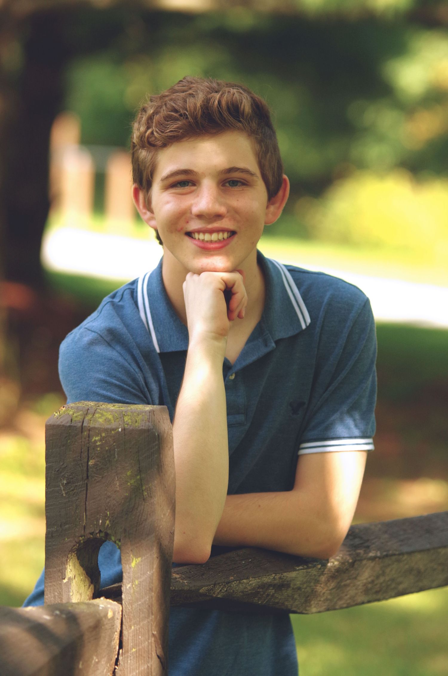 A person in a blue polo shirt leans on a wooden fence post in an outdoor setting.