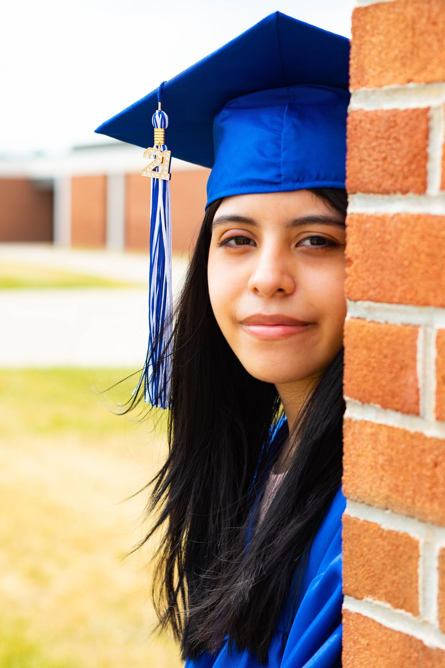 Graduate in blue cap and gown poses against brick wall during commencement.