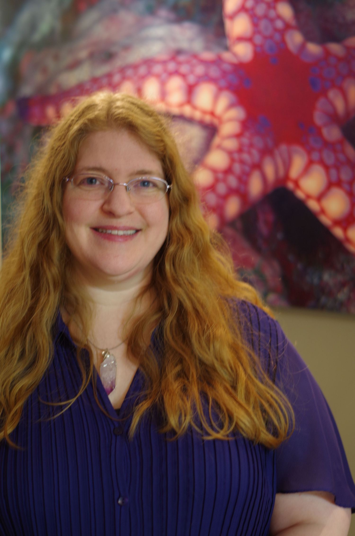 Portrait showing someone with wavy red hair and glasses wearing a deep purple blouse against a decorative backdrop.