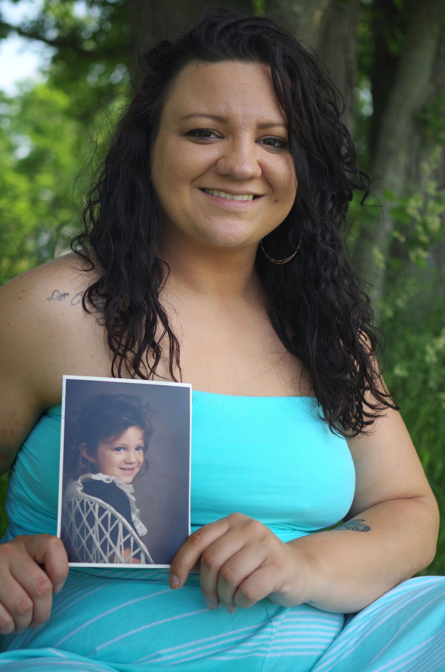 Graduate in turquoise dress holds childhood photo while posing outdoors.
