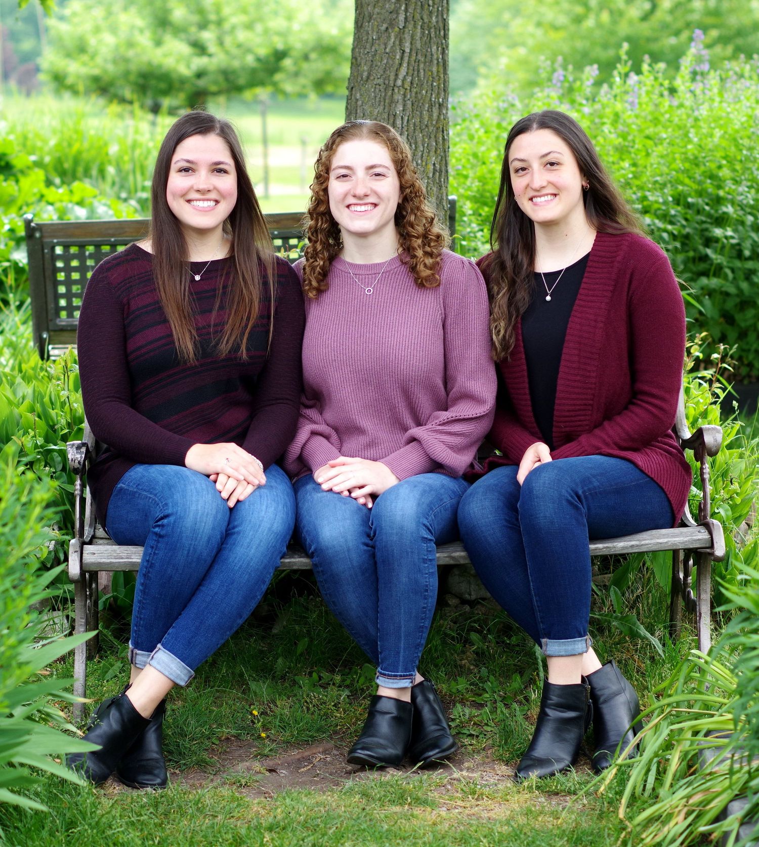 Three young women smiling and sitting together on a wooden bench surrounded by lush green garden foliage.