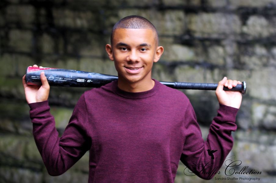 Smiling young man holding a baseball bat over his shoulders, wearing a maroon long-sleeve shirt outdoors.