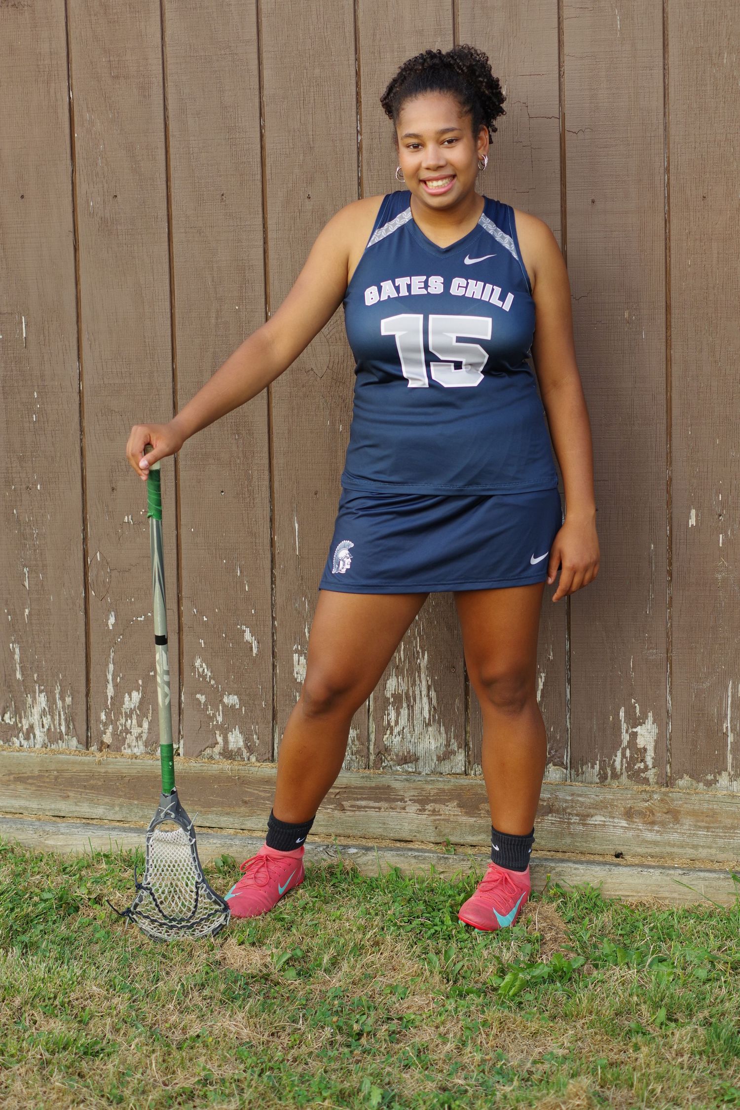 Athlete in navy sports uniform holds lacrosse stick while posing against wooden wall.