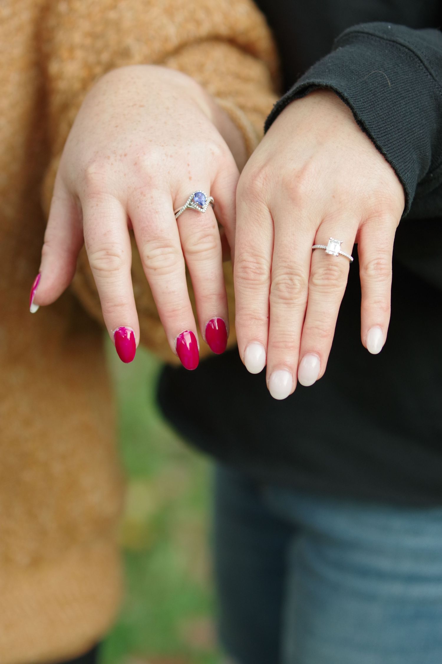 Two women displaying engagement rings, one with red nails and a blue stone ring, the other with nude nails and a diamond ring.