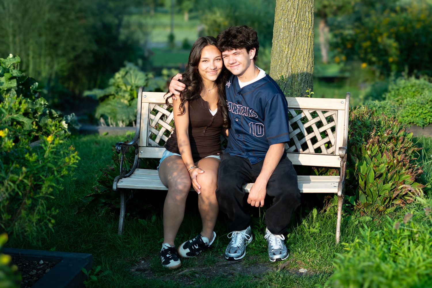 Two teenagers sitting close together on a white garden bench surrounded by lush green plants and trees.