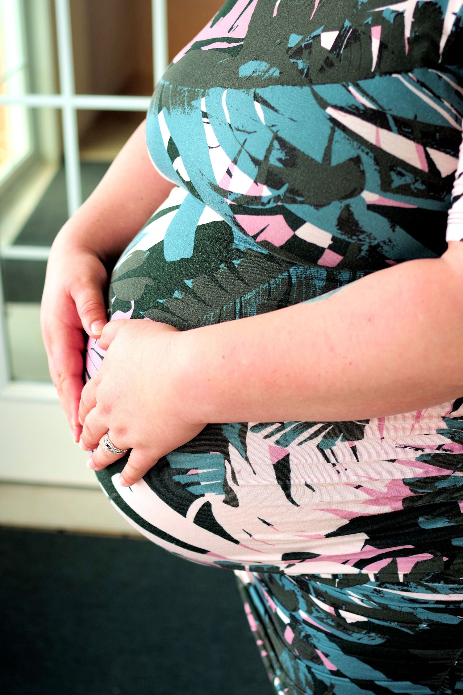 Pregnant woman in floral dress cradling her baby bump with both hands, wearing a ring on her finger.