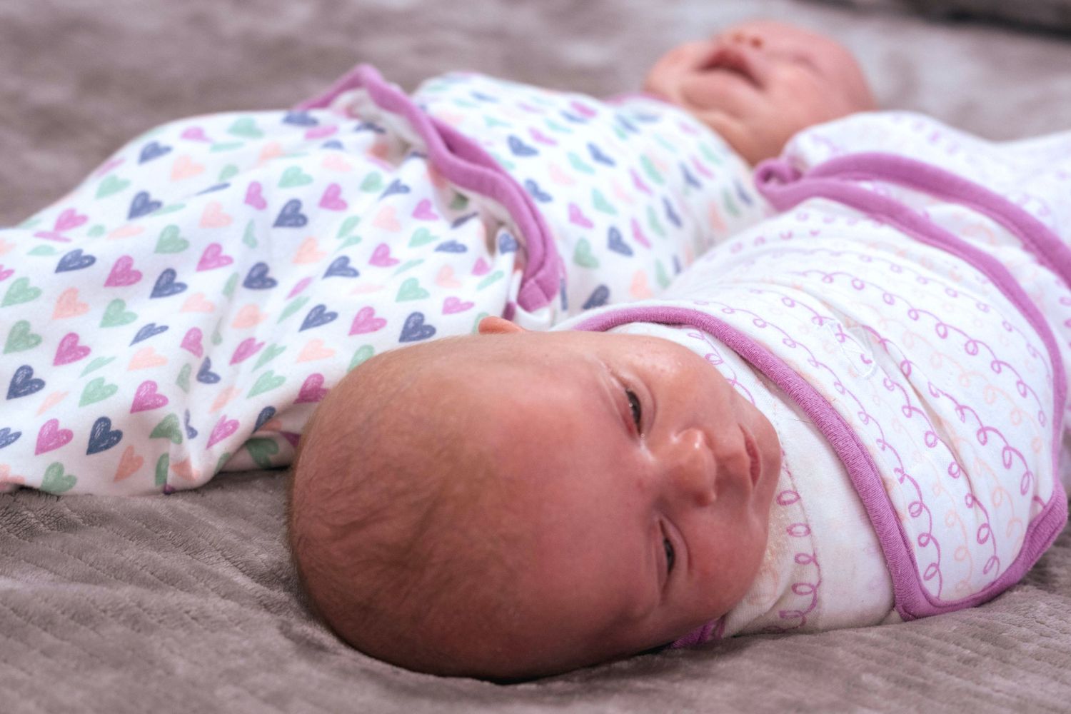Two newborn babies swaddled in heart-patterned and striped wraps lying on a gray surface.