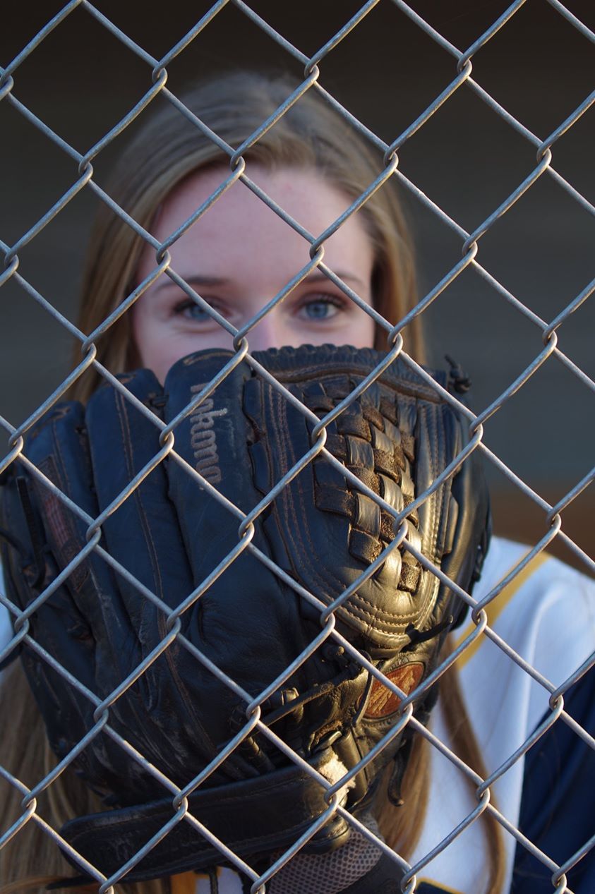 Young female softball player peers through chain-link fence holding a Wilson baseball glove up to her face.