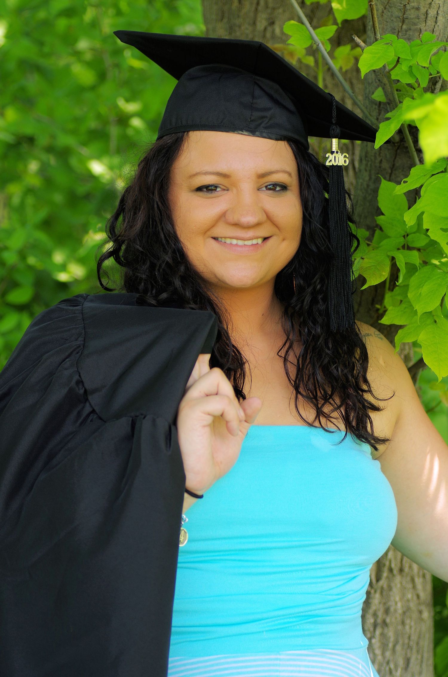 Graduate wearing cap and gown with turquoise dress poses by tree in outdoor setting.