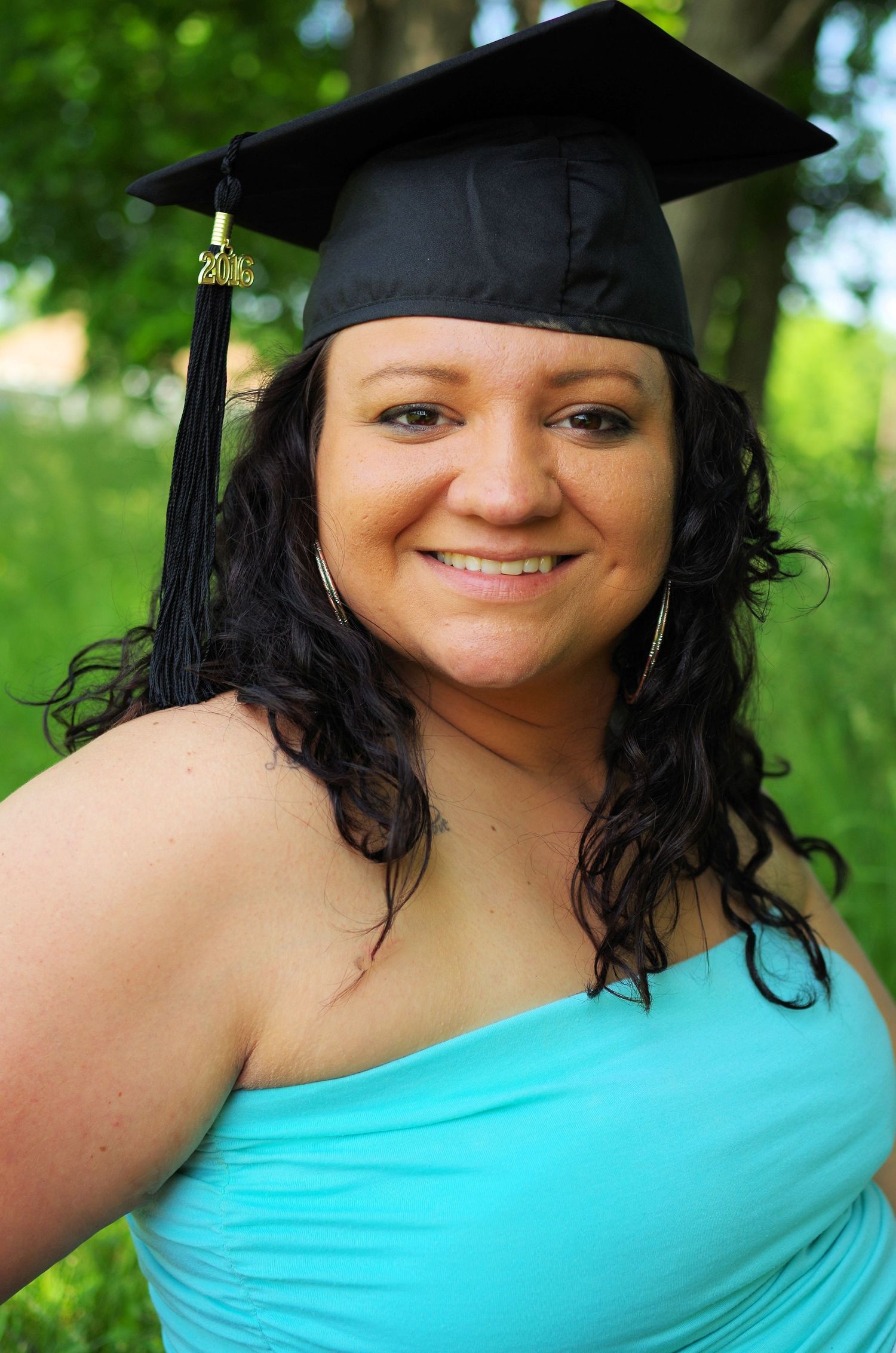 Graduate wearing cap and turquoise dress poses outdoors against green foliage background.