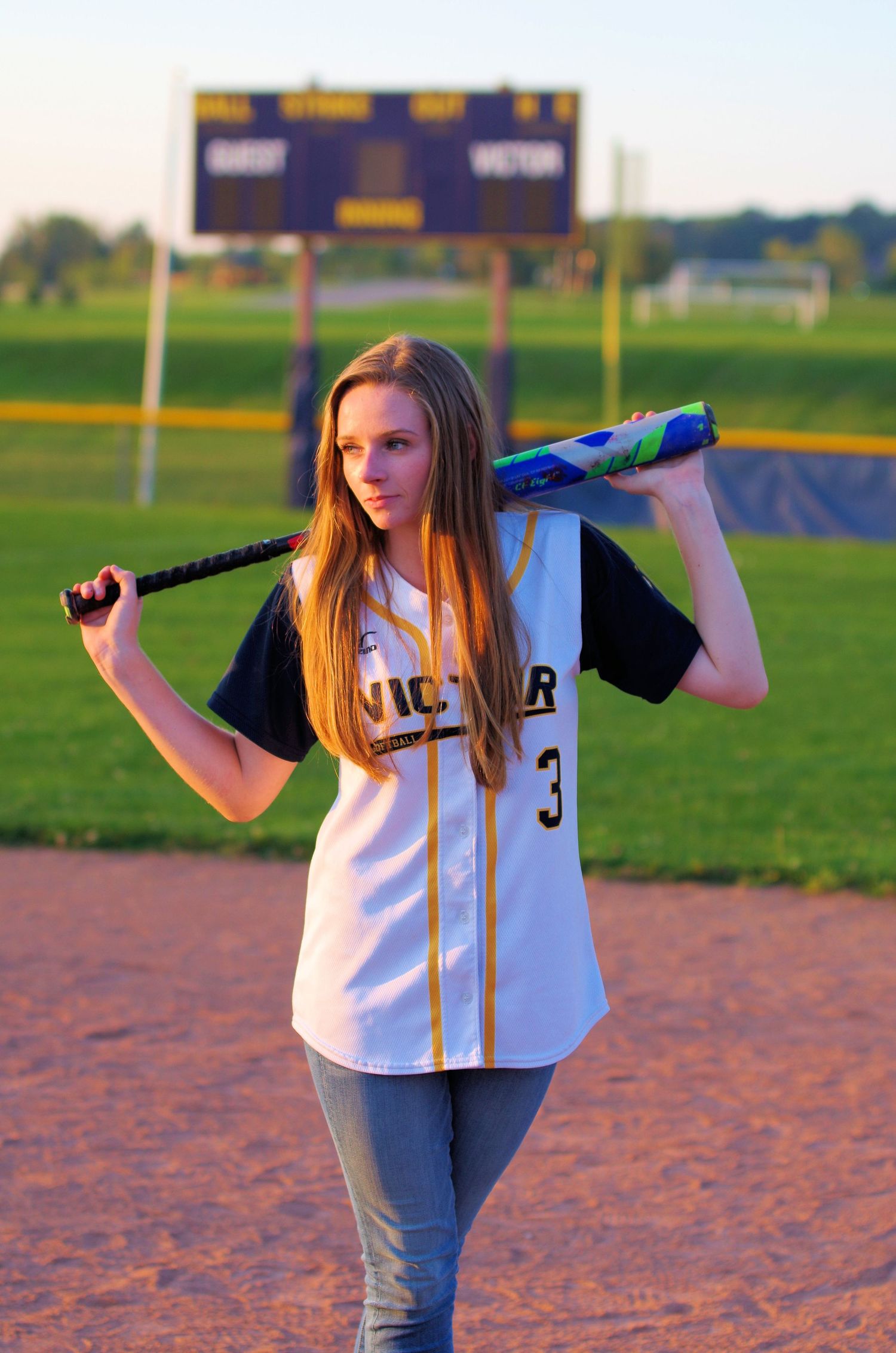 Softball player poses with bat on field with scoreboard visible in background.