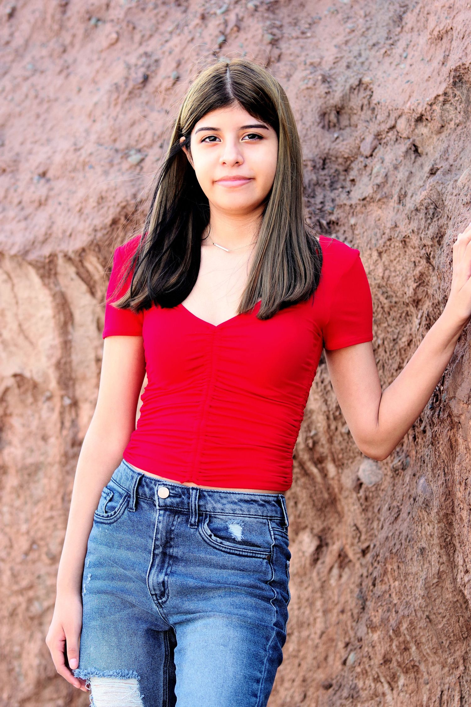 A portrait in a bright red top and jeans standing against a red rock wall.