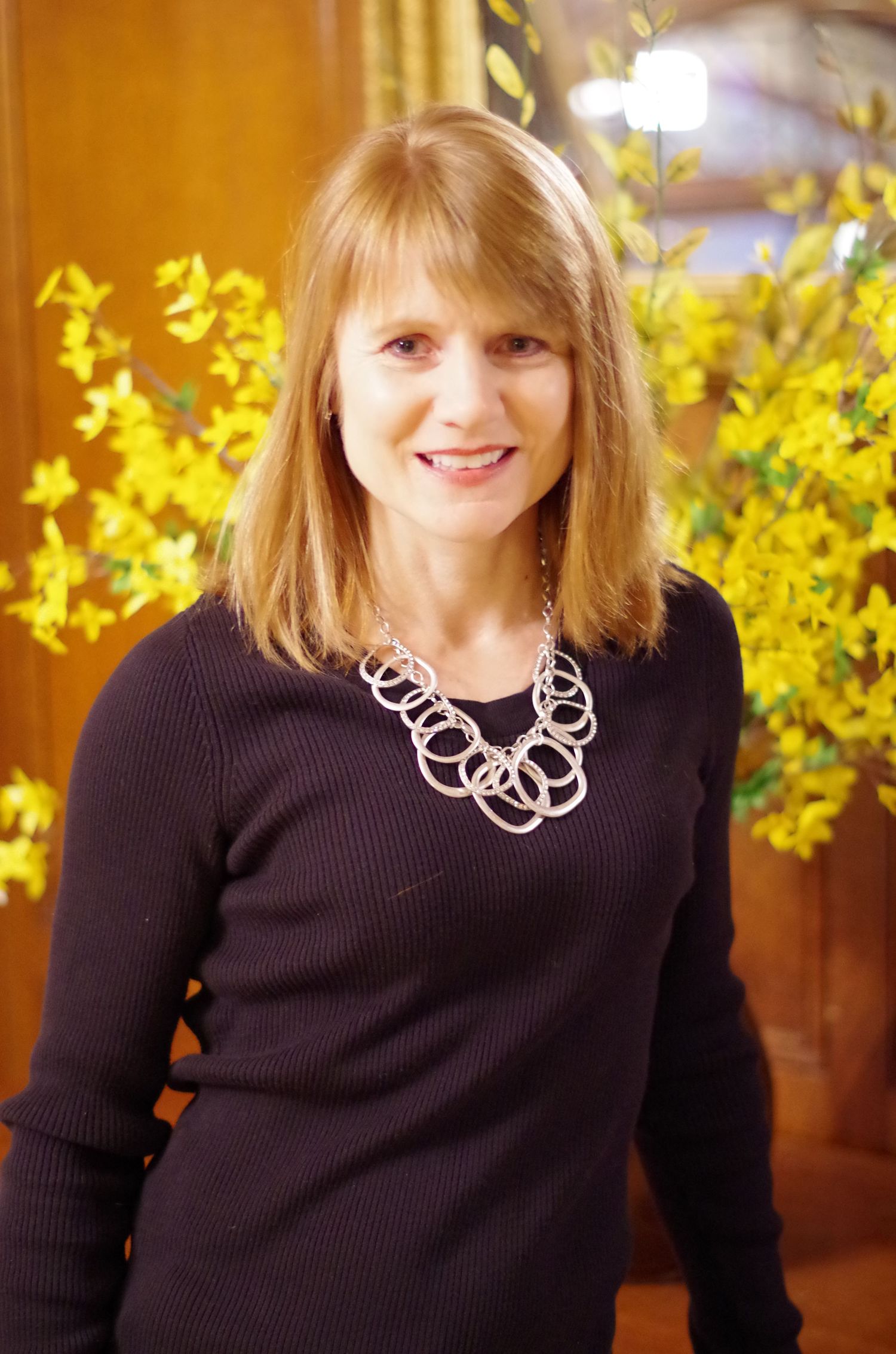 Professional portrait wearing dark sweater and silver chain necklace with yellow forsythia flowers in background.