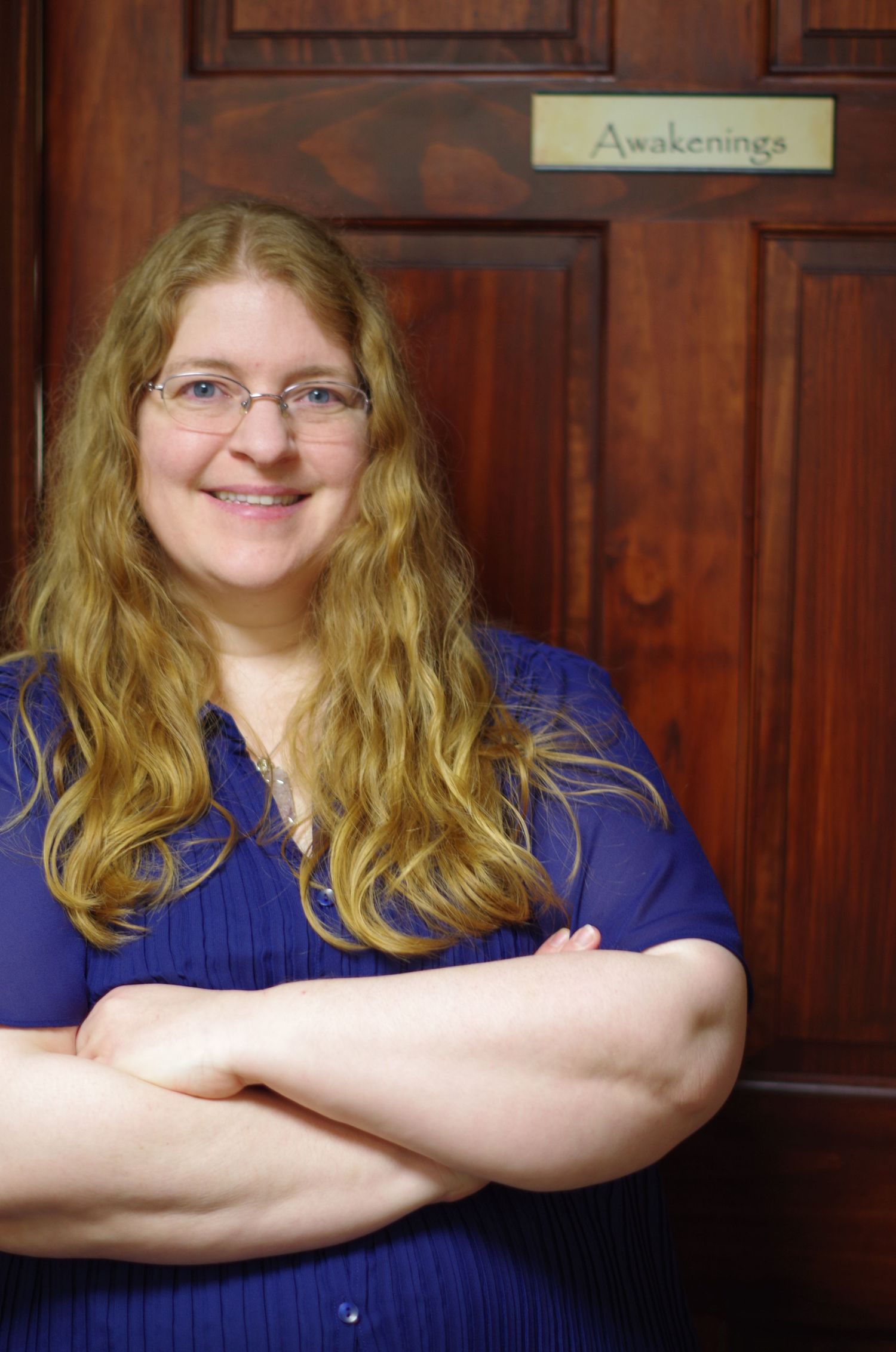 Portrait of someone with long wavy hair in a navy button-down shirt standing by a wooden door labeled Awakenings.