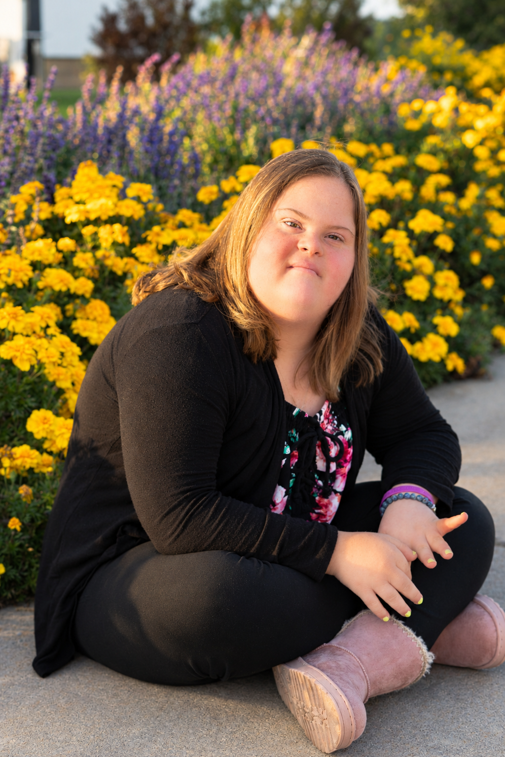 Young woman sitting cross-legged in front of vibrant yellow and purple flowers, smiling warmly outdoors.
