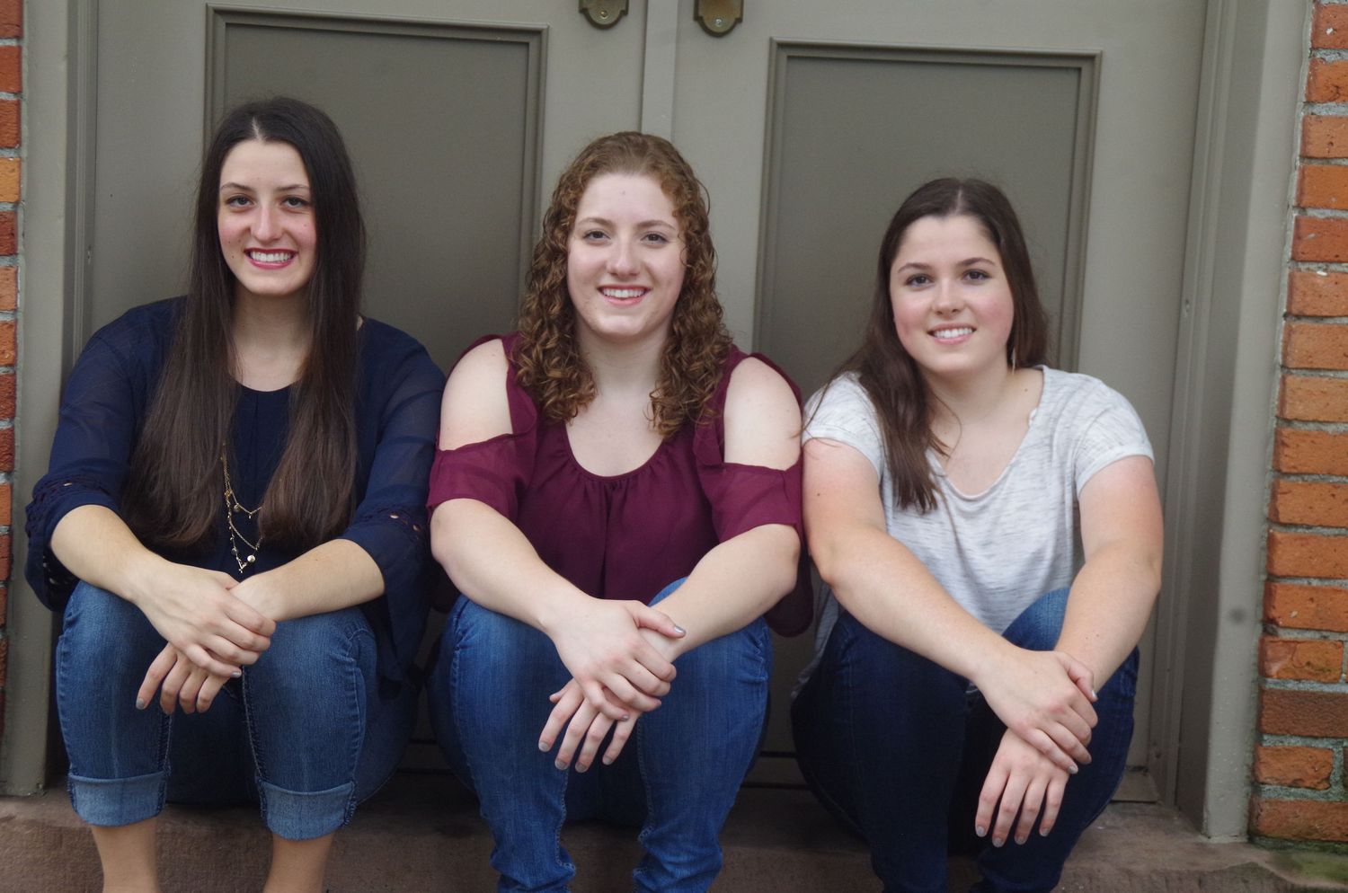 Group of students sitting together on steps in front of grey door with brick walls.