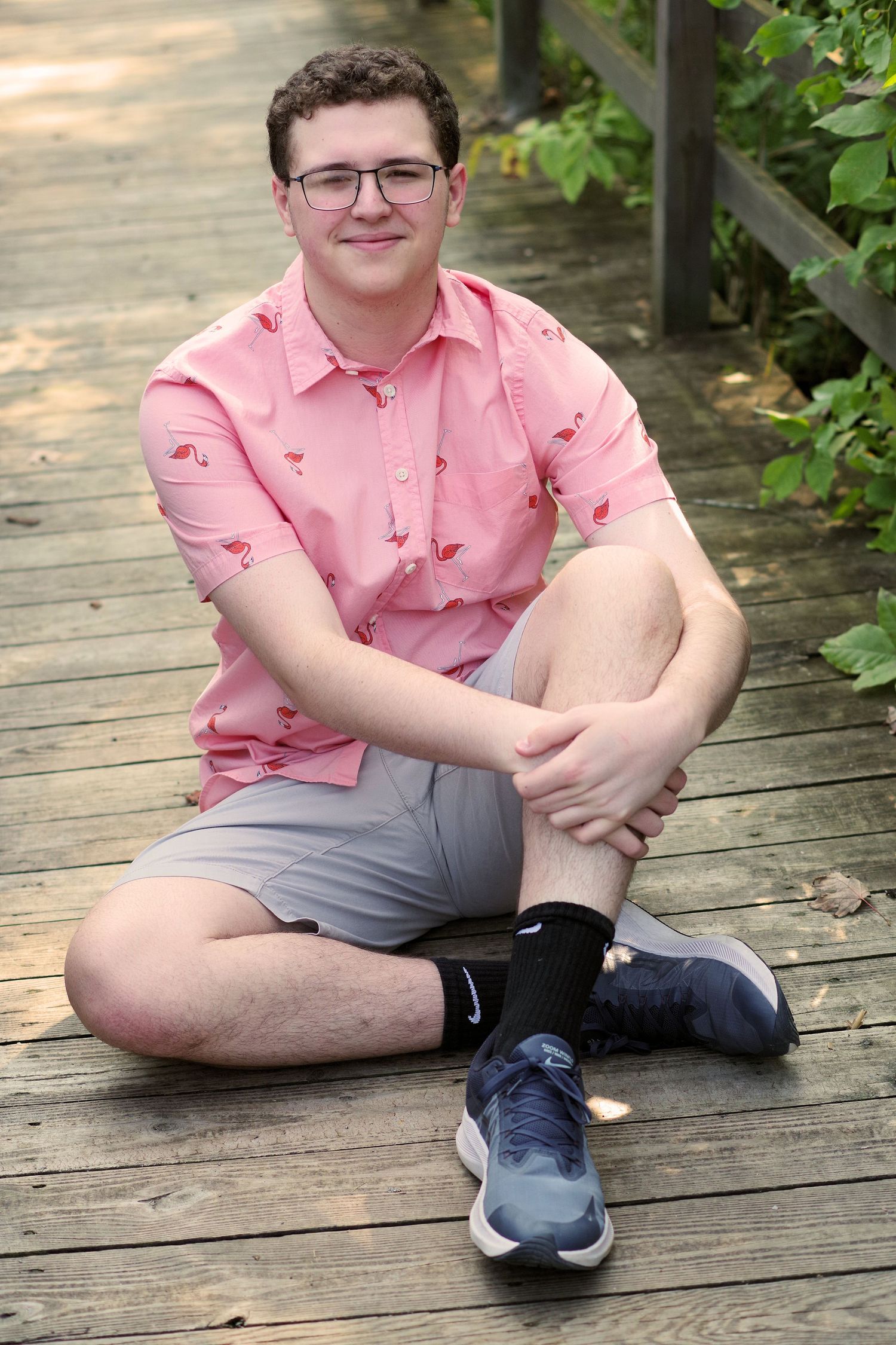 A seated figure in a pink patterned shirt and gray shorts on a wooden boardwalk surrounded by greenery.