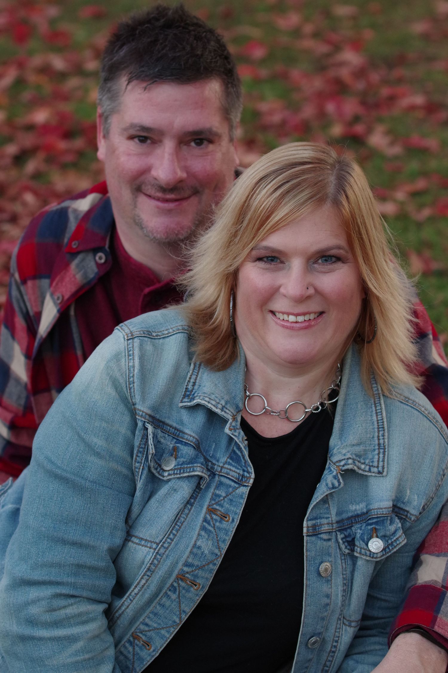 Couple in casual attire smiling for portrait with fall foliage background.