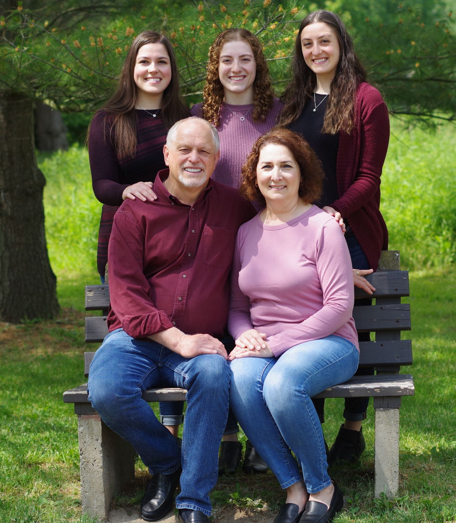 Family of five posing outdoors on a wooden bench, parents seated with three daughters standing behind them.