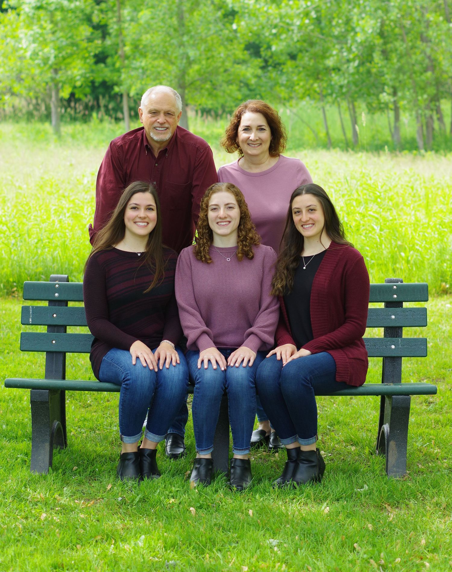 Family of five posing outdoors on a green bench, with lush green trees in the background on a sunny day.
