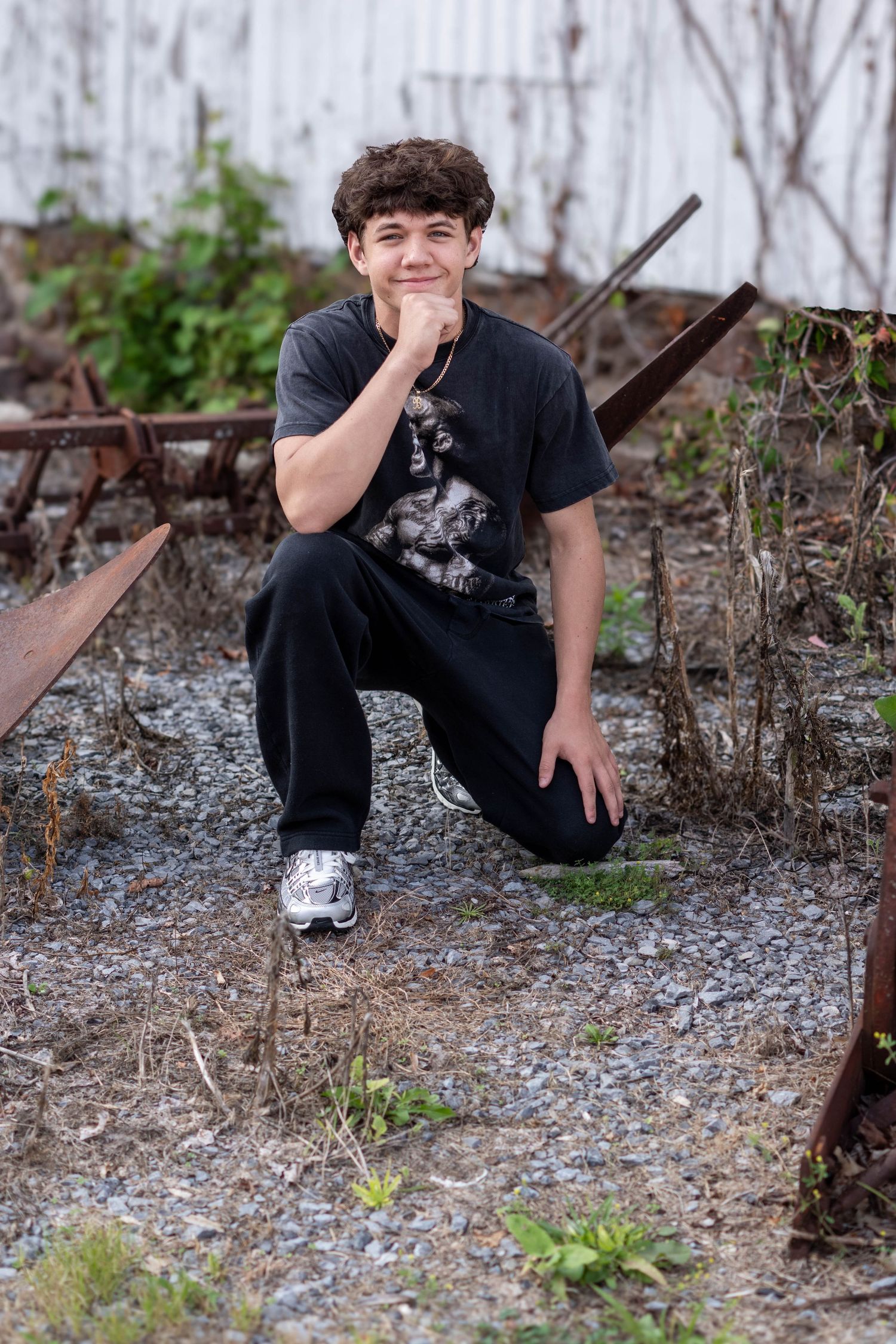 Teen boy in black outfit crouching on gravel near rusty metal rails, smiling confidently in an urban decay setting.