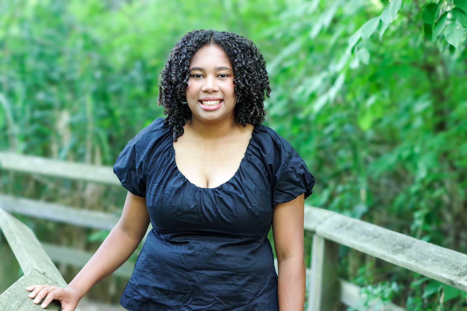 Someone wearing a black top smiles while sitting on a wooden railing with lush green foliage in the background.