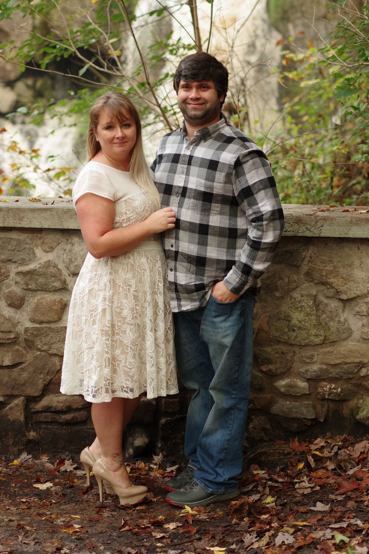 Romantic couple posing by stone wall in garden setting wearing casual attire.