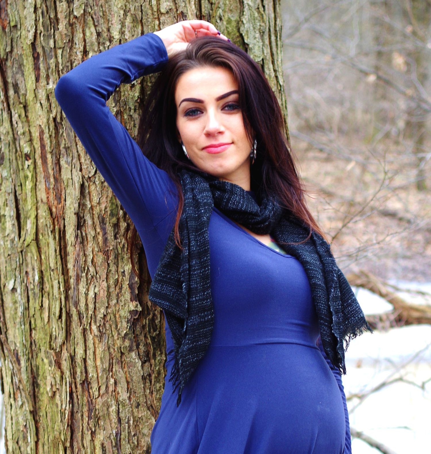Maternity photo of woman in navy blue dress posed against snowy forest backdrop.