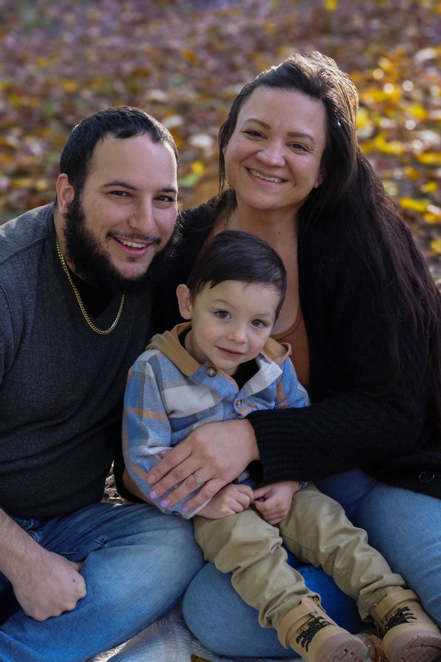 Small family seated together among fallen autumn leaves during outdoor photo session.
