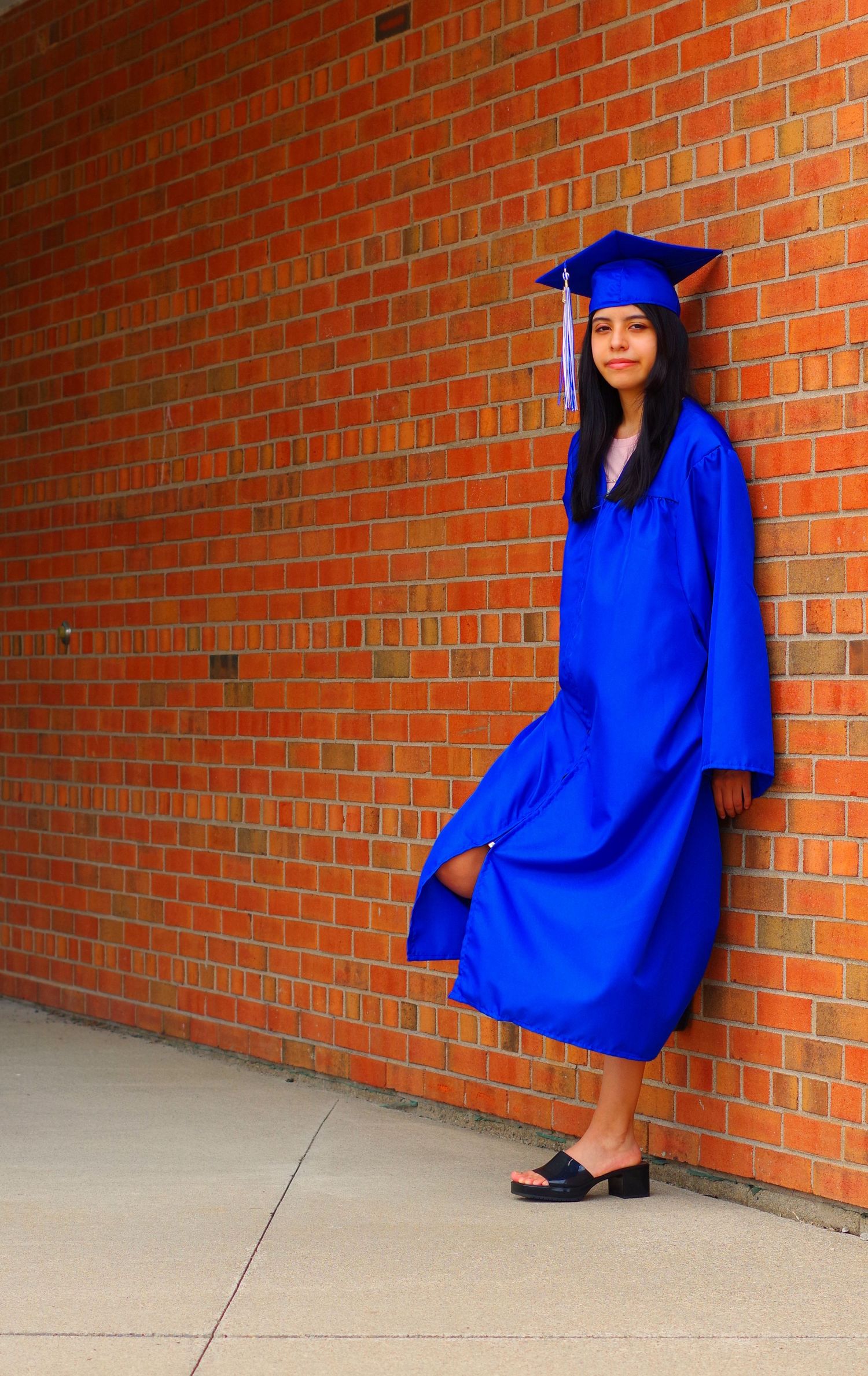 Full-length graduation photo showing graduate seated against red brick wall.