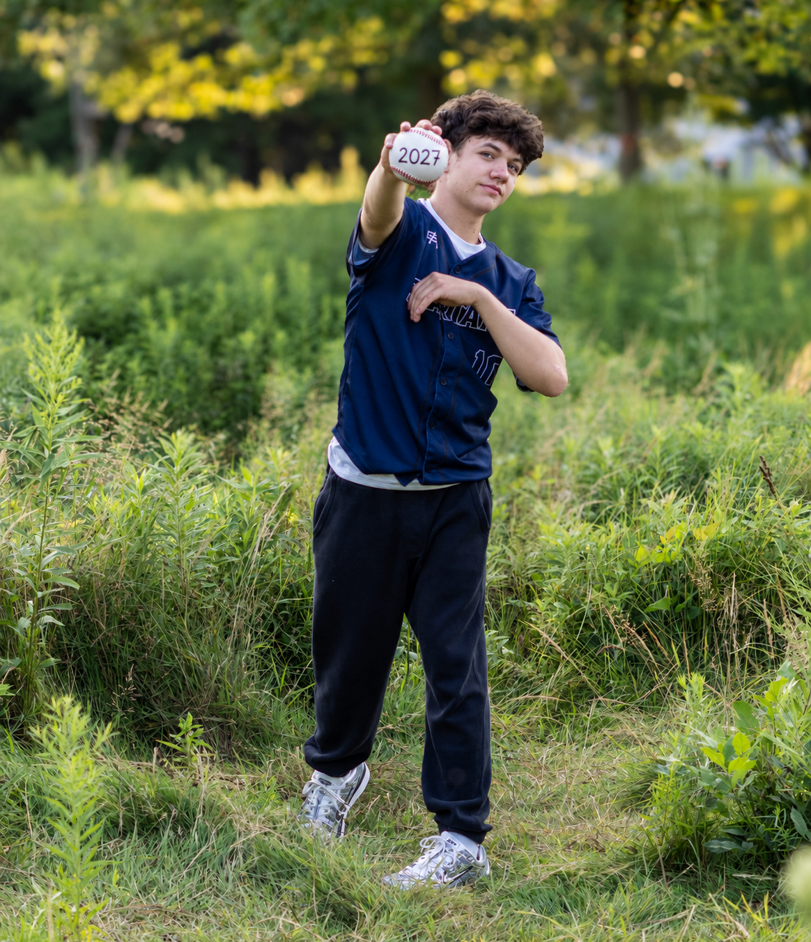 Teen boy in navy jersey holding a ball outdoors in a lush green field during a sunny day.