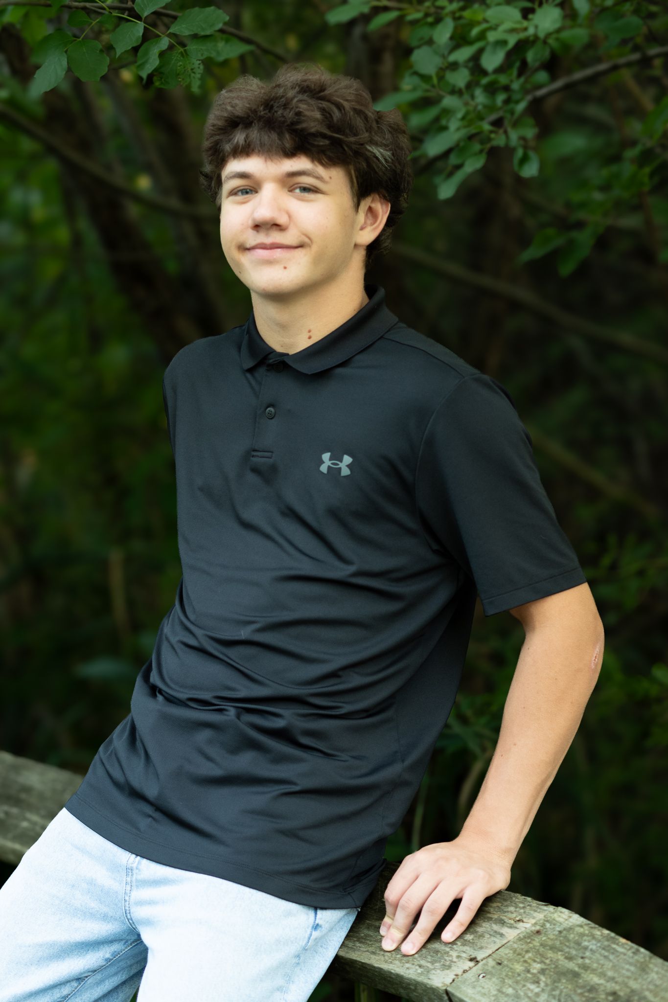Young man in black Under Armour polo shirt leaning on wooden railing outdoors with green trees background.