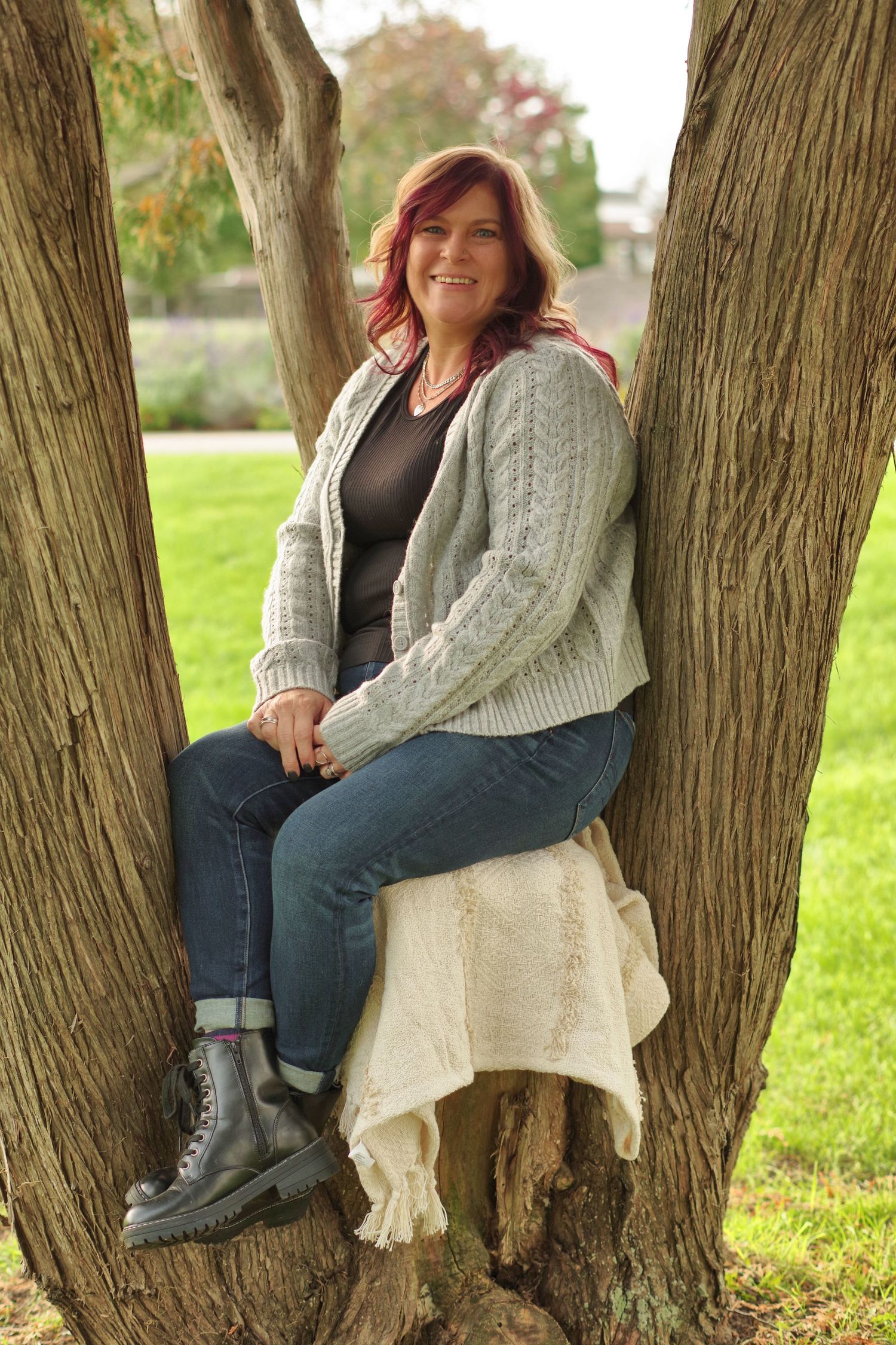 A smiling redhead woman sits in a tree fork wearing a gray cardigan, jeans, and boots in a green park.