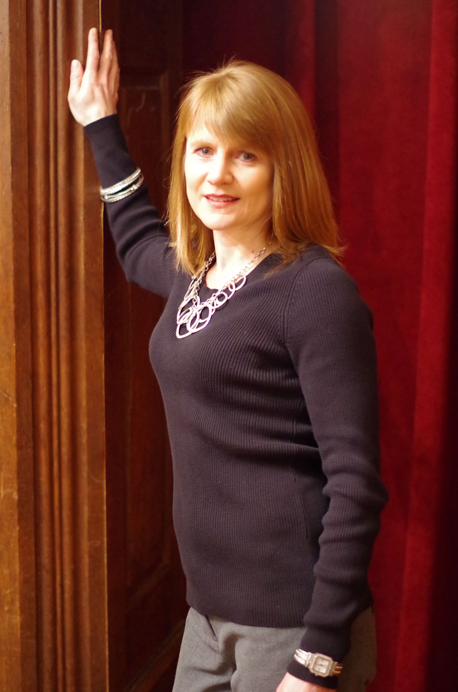 Side view portrait with raised hand wearing black sweater and statement necklace against red curtain.