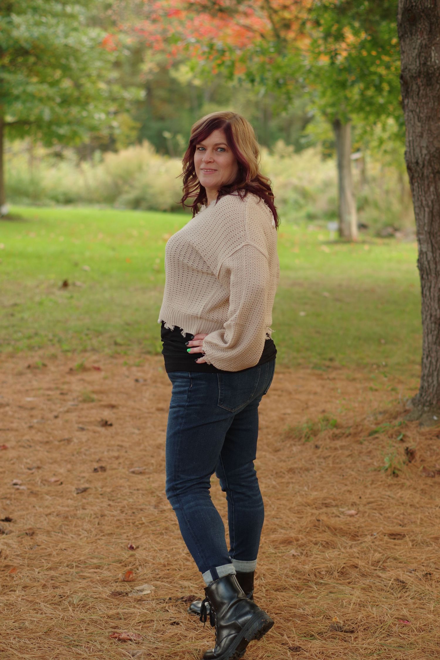 Person in cream sweater and boots poses on autumn path with fall colors in background.