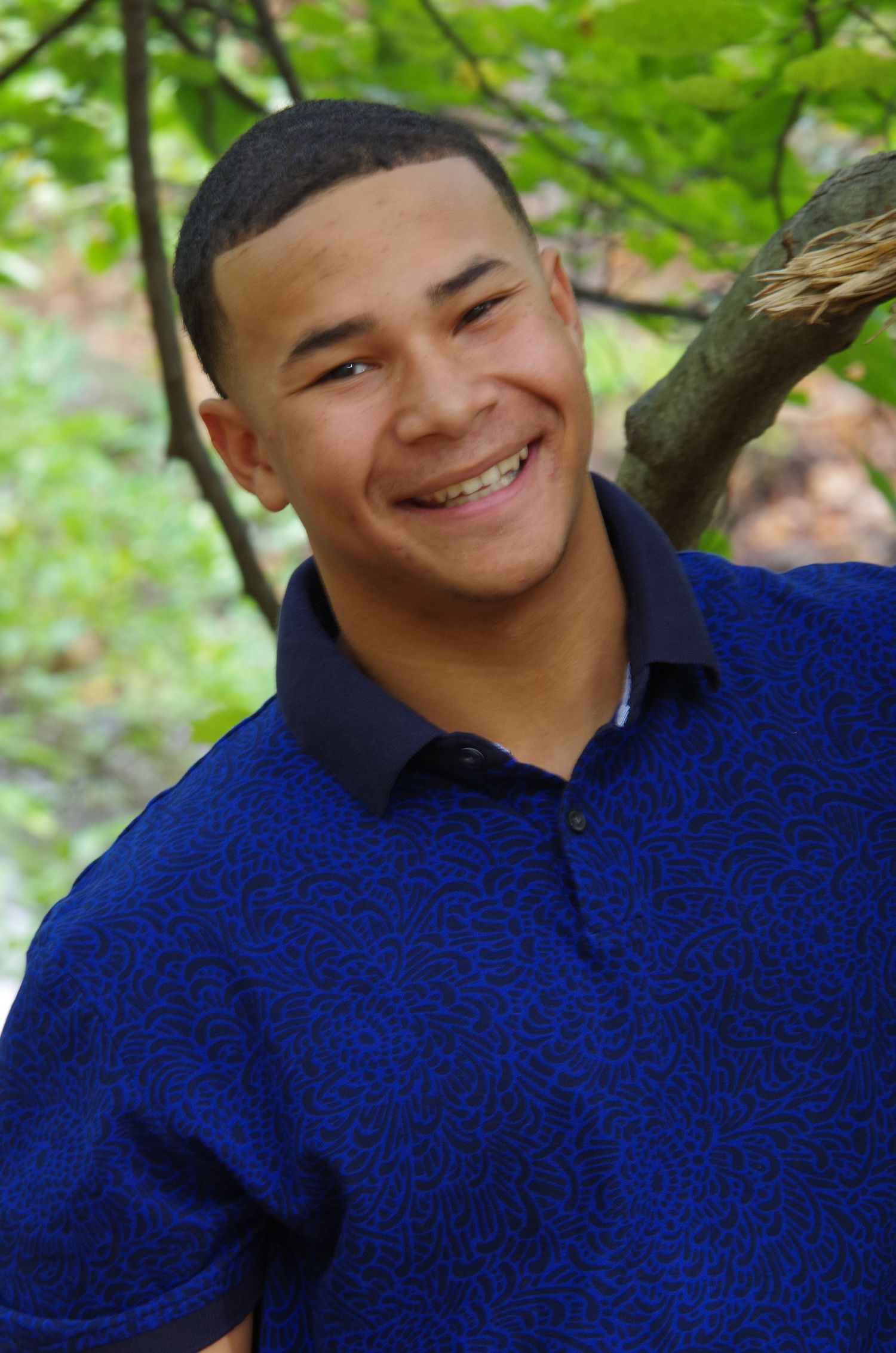 A person wearing a royal blue polo shirt smiling against a green leafy background.