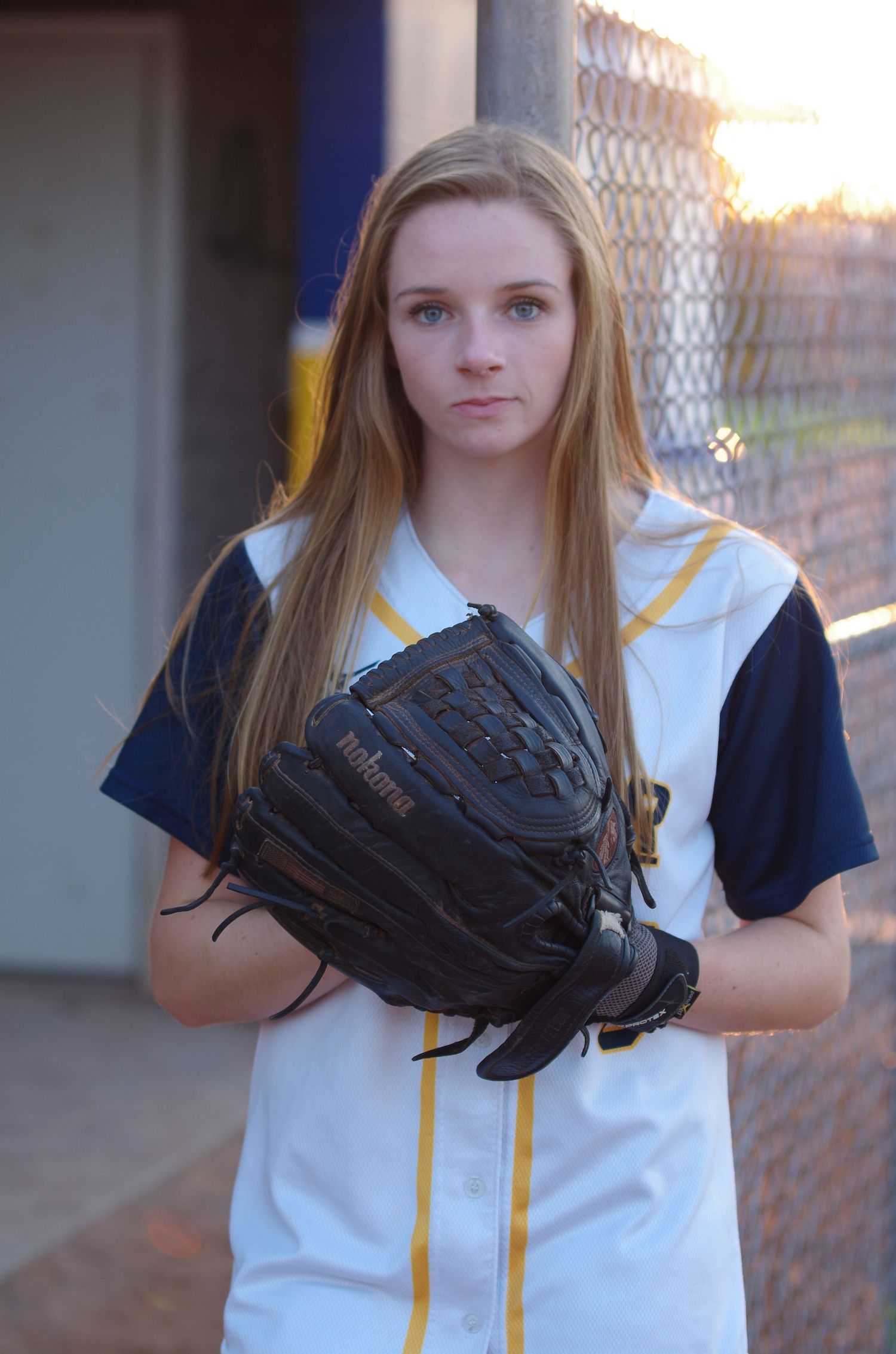 Baseball player in white and navy uniform holds glove against fence background.