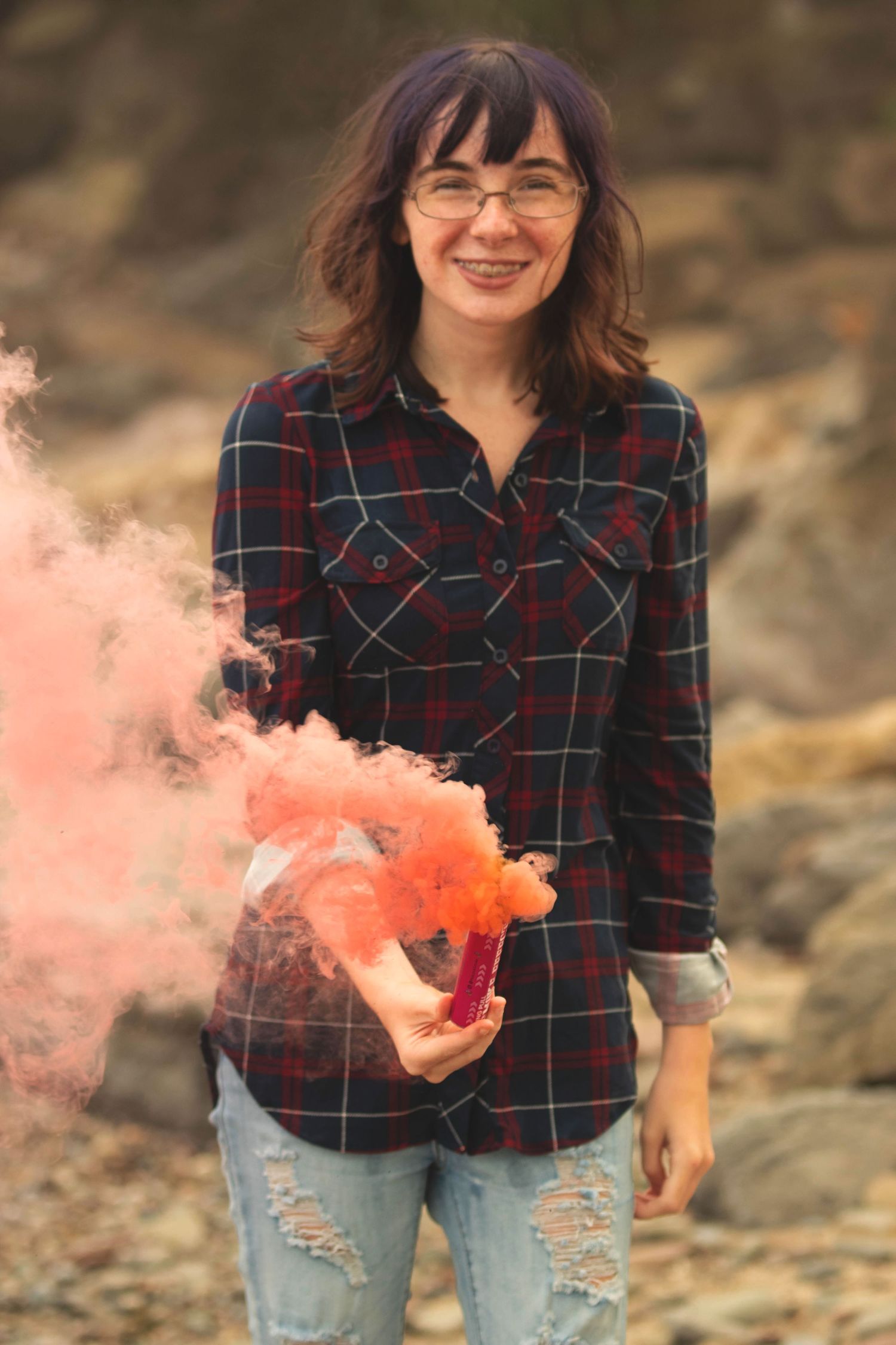 Person holding orange smoke bombs while posing on rocky terrain in plaid shirt and distressed jeans.