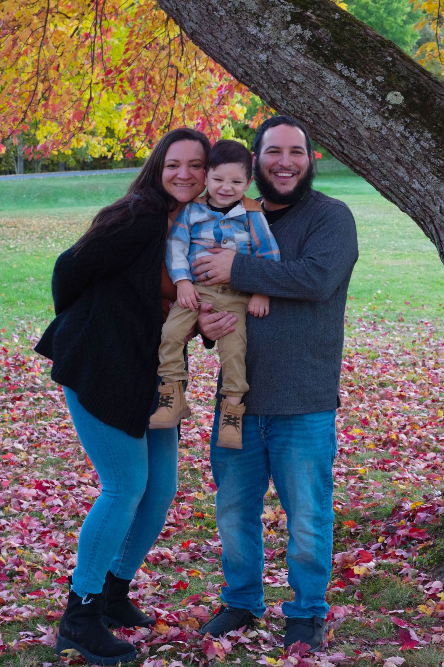 Family stands together under large tree branch with colorful fall foliage in background.